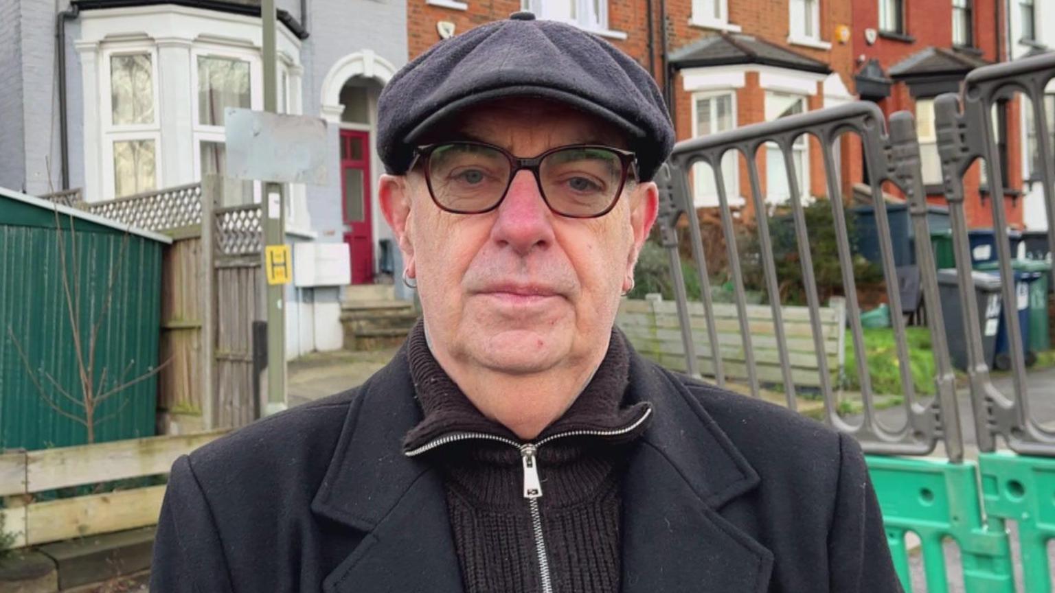 A man in a flat cap, black coat and zip jumper with glasses looks at the camera. Behind him are houses and temporary works fencing.