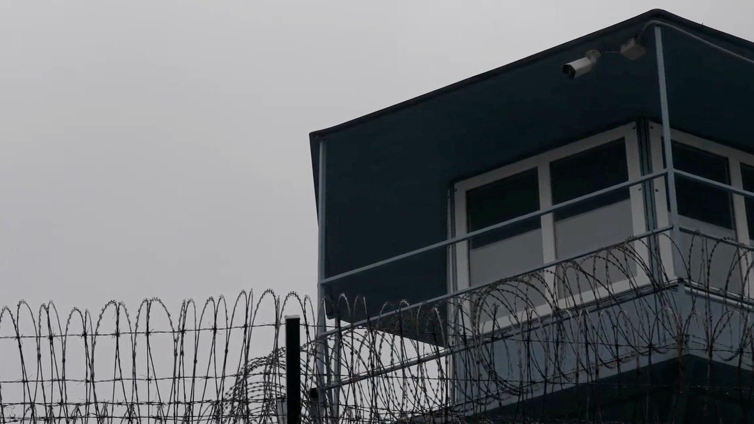 A picture of a grey watchtower with white frames around the windows. Barbed wire is strung along the top of the nearby fence at the bottom of the picture. CCTV cameras can be seen pointing into the prison.