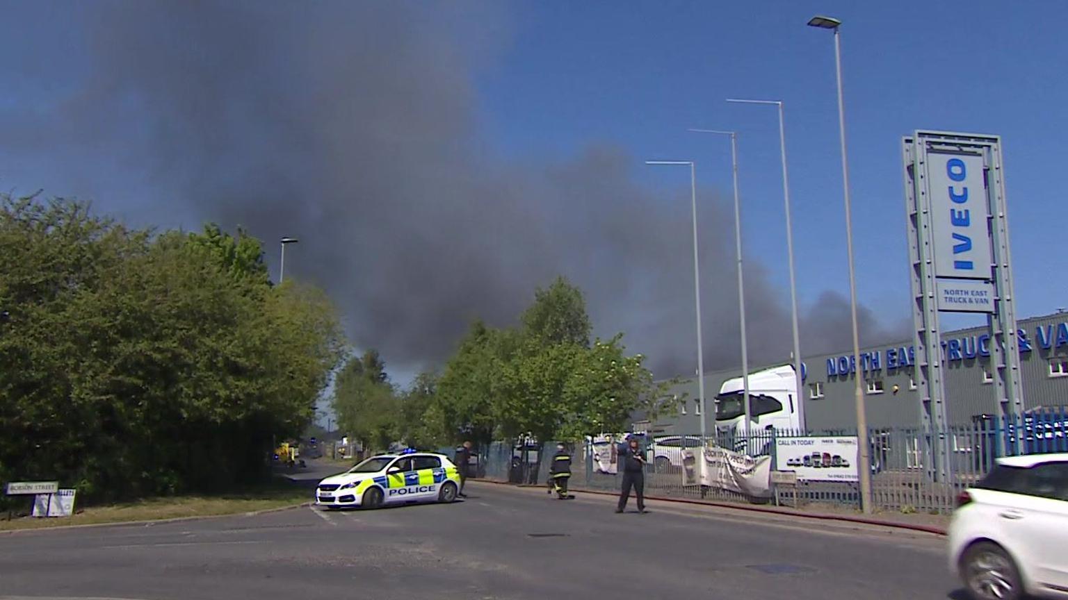 A large black smoke plume can be seen blowing over an IVECO garage with a parked lorry behind a large fence. A police car is parked across the street and a firefighter is walking in the direction of the smoke.