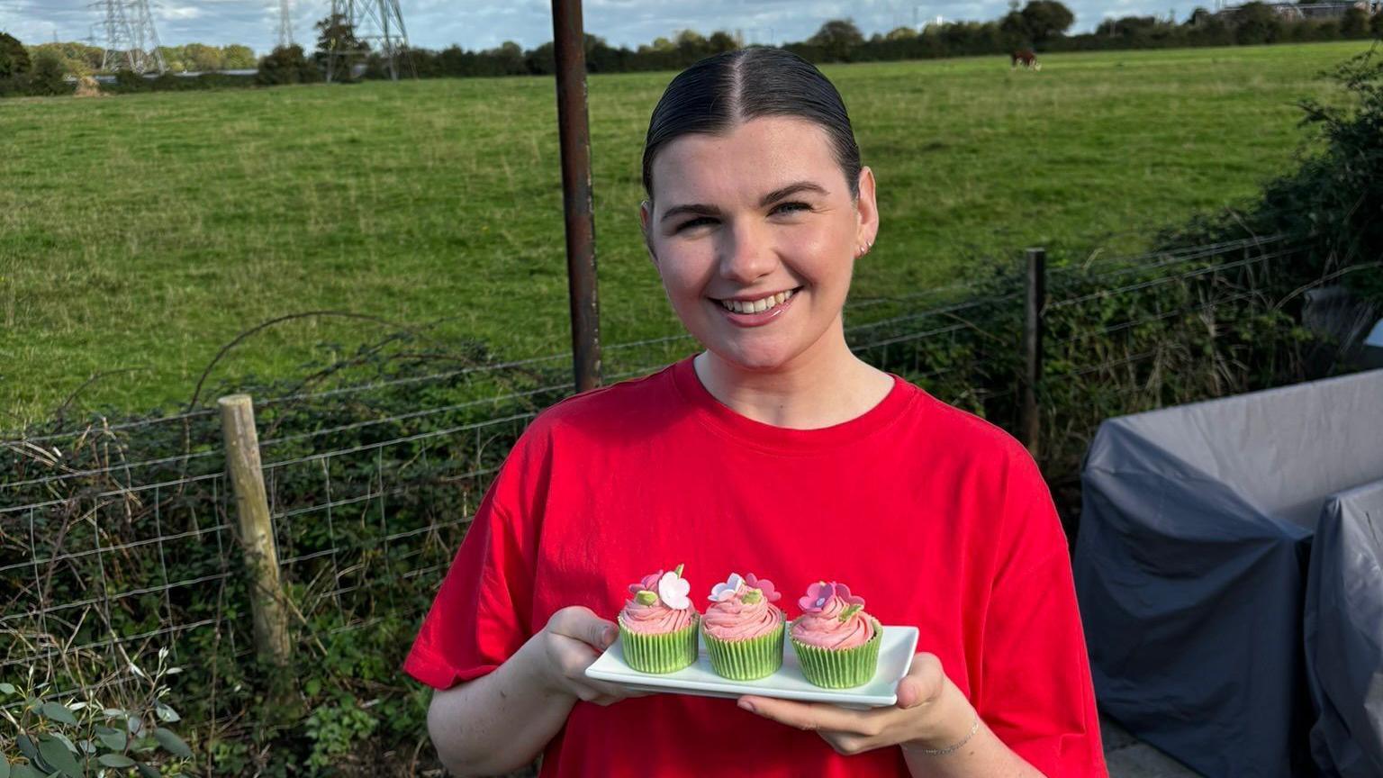 Katy Davies smiling holding a tray of cupcakes for the homeless, she wears a red t-shirt and has dark brown hair slicked back into a bun. 