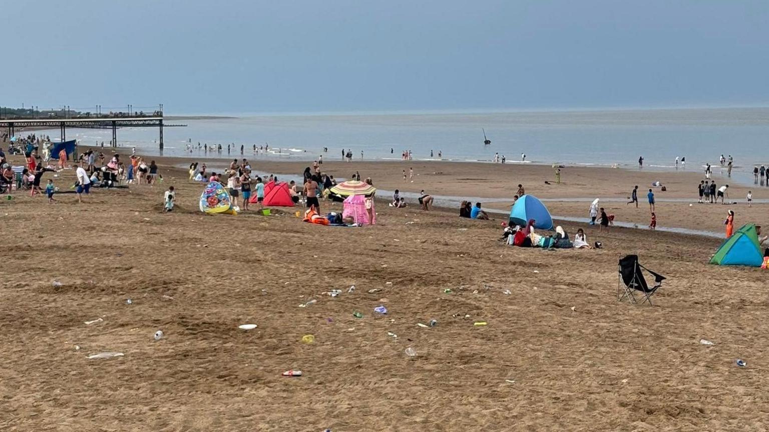 Sandy beach with litter on it. There are people and tents and a folding chair. There are pylons in the distance There is a sea.