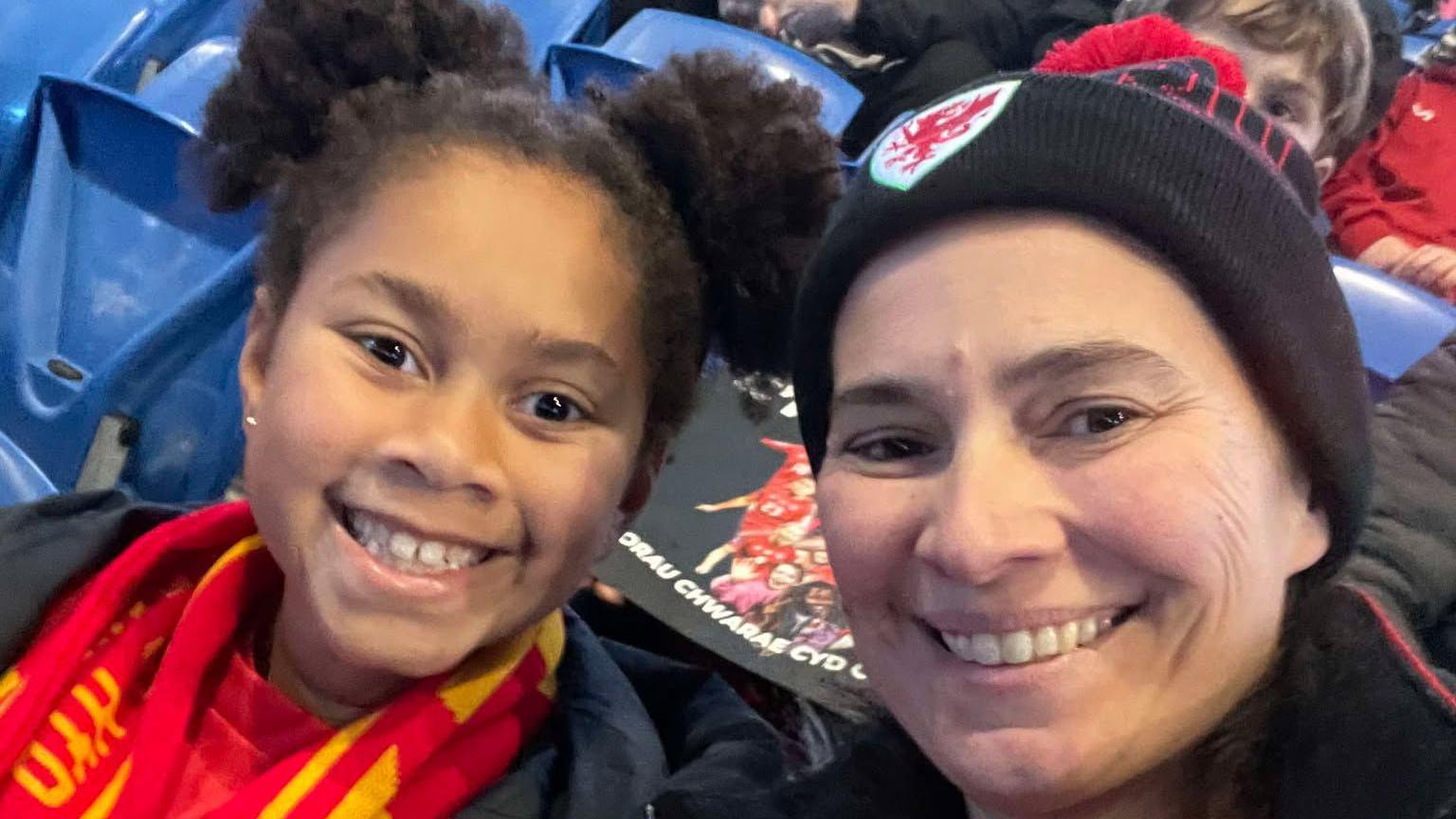 45-year-old Marisa (right) and her daughter Yasmin (left) smile taking a selfie on the sides in a stadium. 