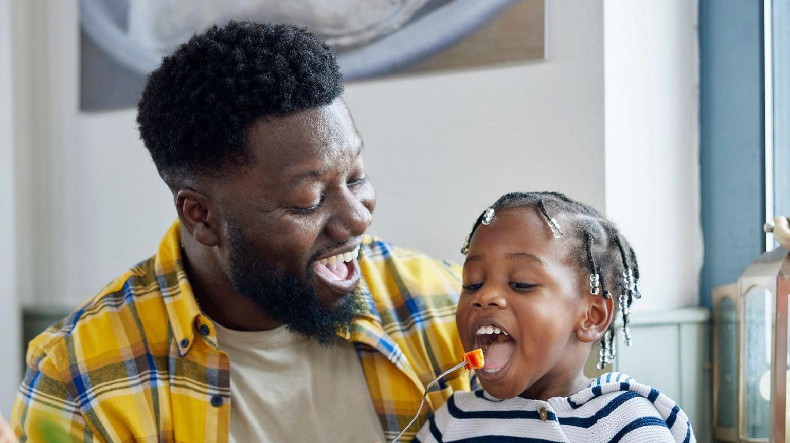 A man feeds a child a piece of fruit on a fork as they both laugh and smile. 