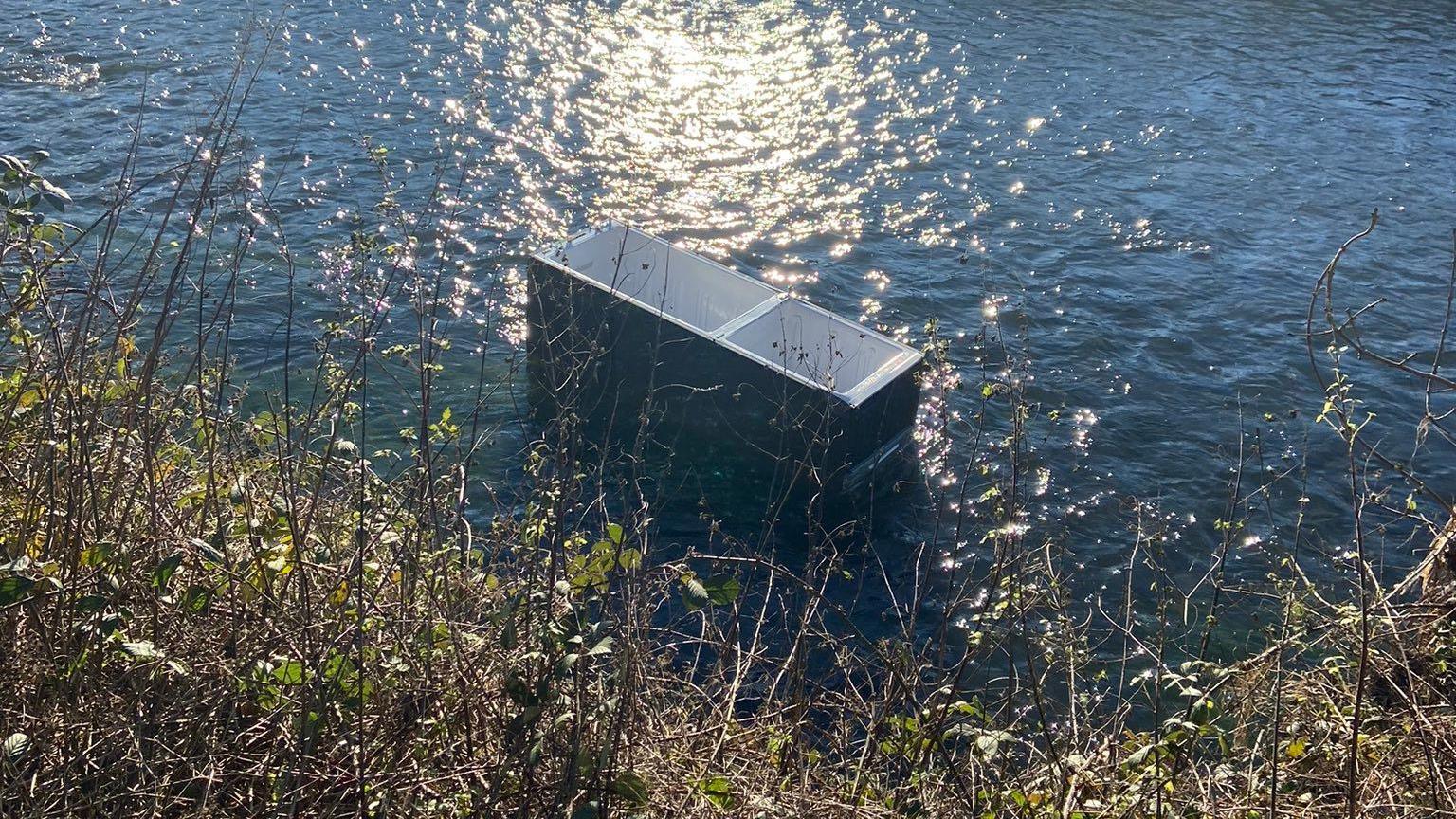 A dark grey fridge freezer is visible in a canal with bright blue water, which shines as the sun reflects off it