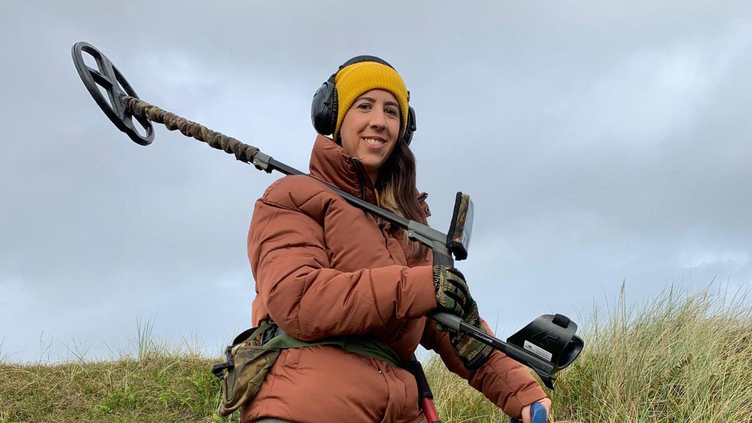 Woman in orange coat, yellow hat and headphones holds a metal detector over her shoulder. She smiles at the camera, army print gloves on and matching bag around her waist. She stands on grass, the sky is overcast.