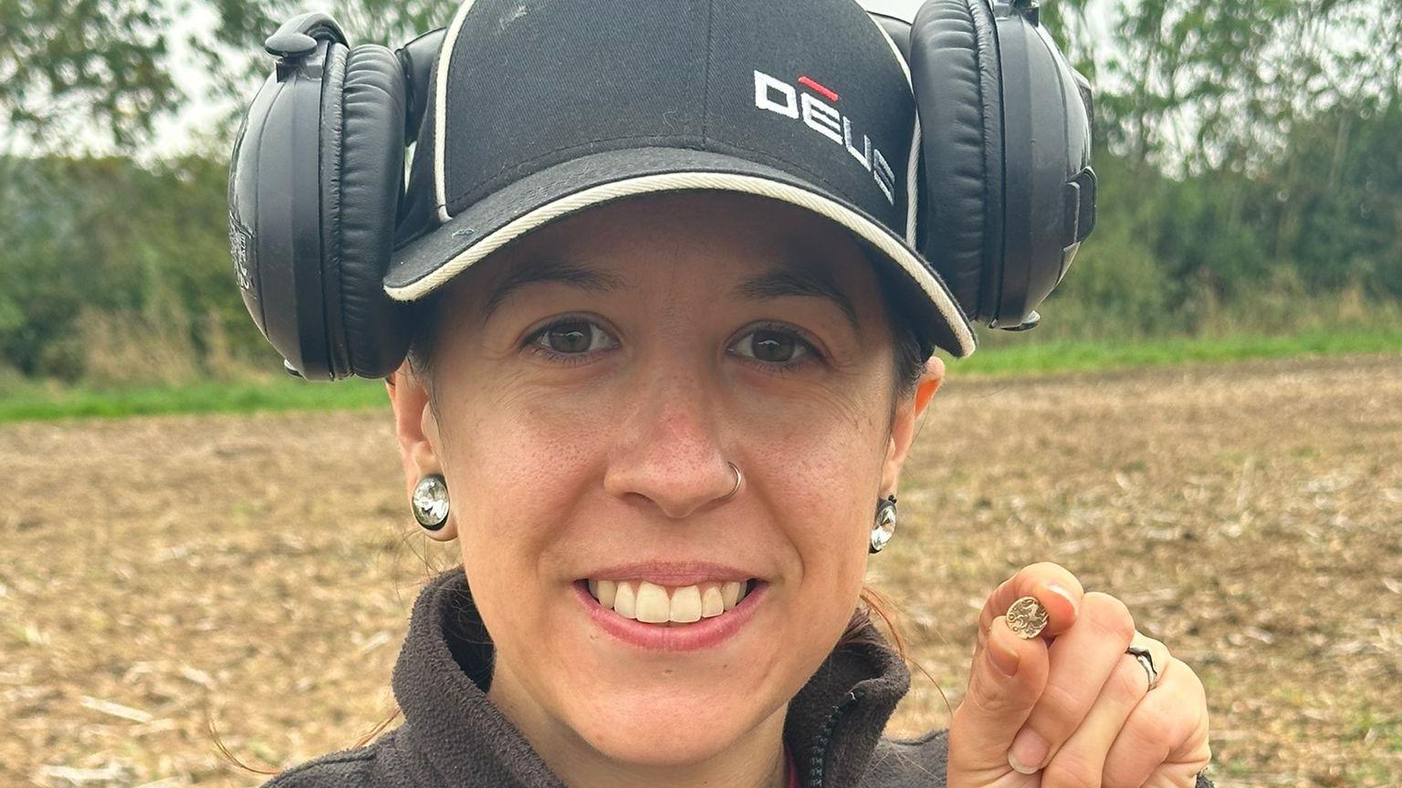 A smiling woman holds up a tiny coin to the camera in a head and shoulders shot. She's wearing a dark fleece, black baseball cap and large black headphones