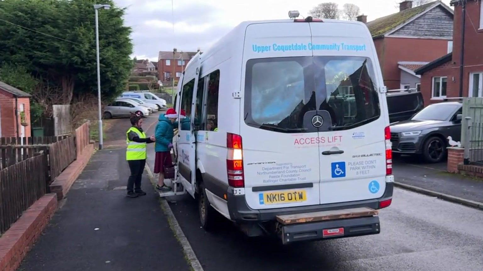 A minibus is parked up and an elderly lady wearing a Santa hat is being helped on board by a woman wearing a yellow hi-vis jacket.