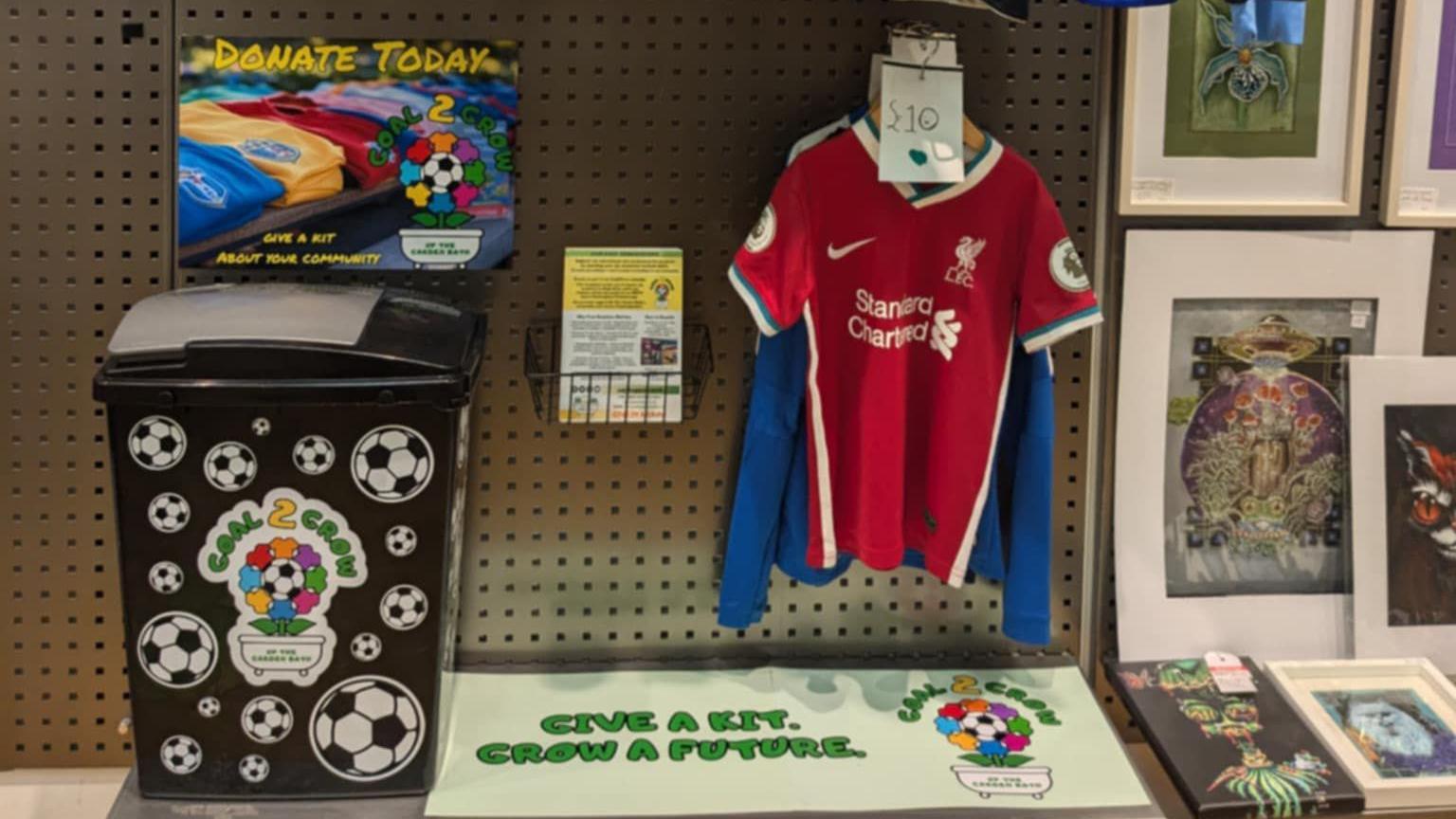 Football shirts, including a red Liverpool jersey, are hanging on a display inside. There is a green sign which says "Give a kit. Grow a future".