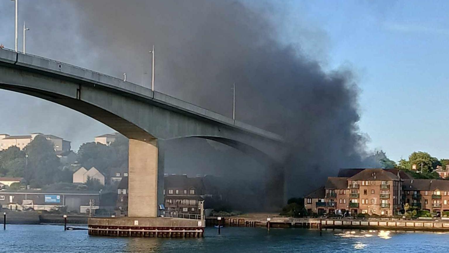 Grey smoke coming from the bottom of a concrete bridge. There is water under the bridge.