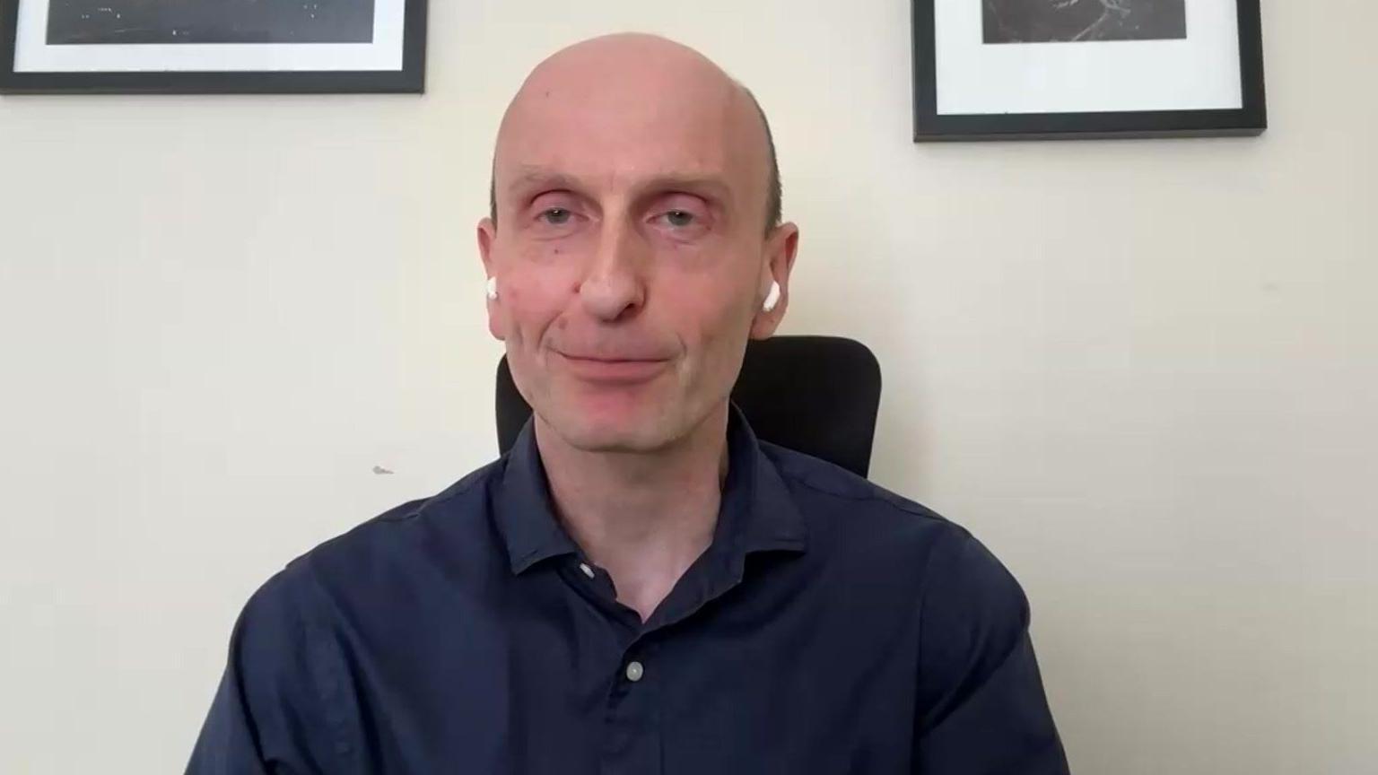 Robert Wilson looking at the camera wearing a navy blue shirt. He is sat in a black chair and the wall behind him is white, with two frames above him
