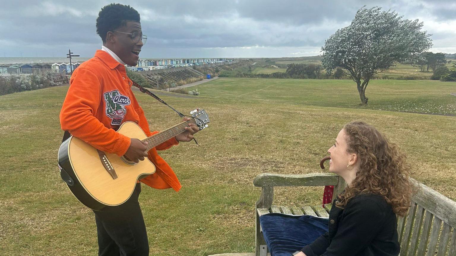 Jordan Benjamin playing a guitar on the Greensward at Frinton-on-Sea. He is looking at Amy Trigg, who is sat on a bench in front of him and smiling.