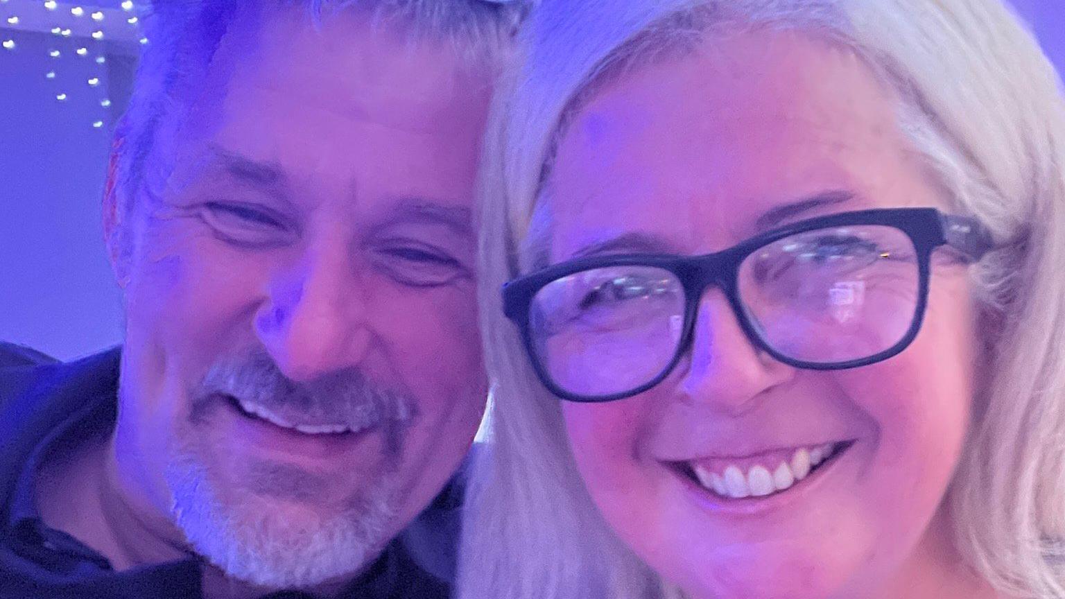 A selfie of Roz on the right and Tim on the left. She has shoulder-length white-blond hair and is wearing black-rimmed glasses. He has grey hair and a short grey goatee and moustache.