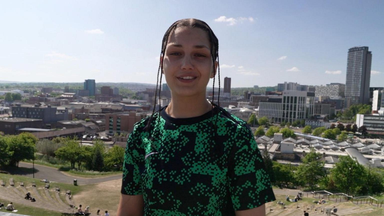 AJ is stood proud in front of the Sheffield landscape. She is wearing a black and green pattered T-shirt and her hair is in braids.