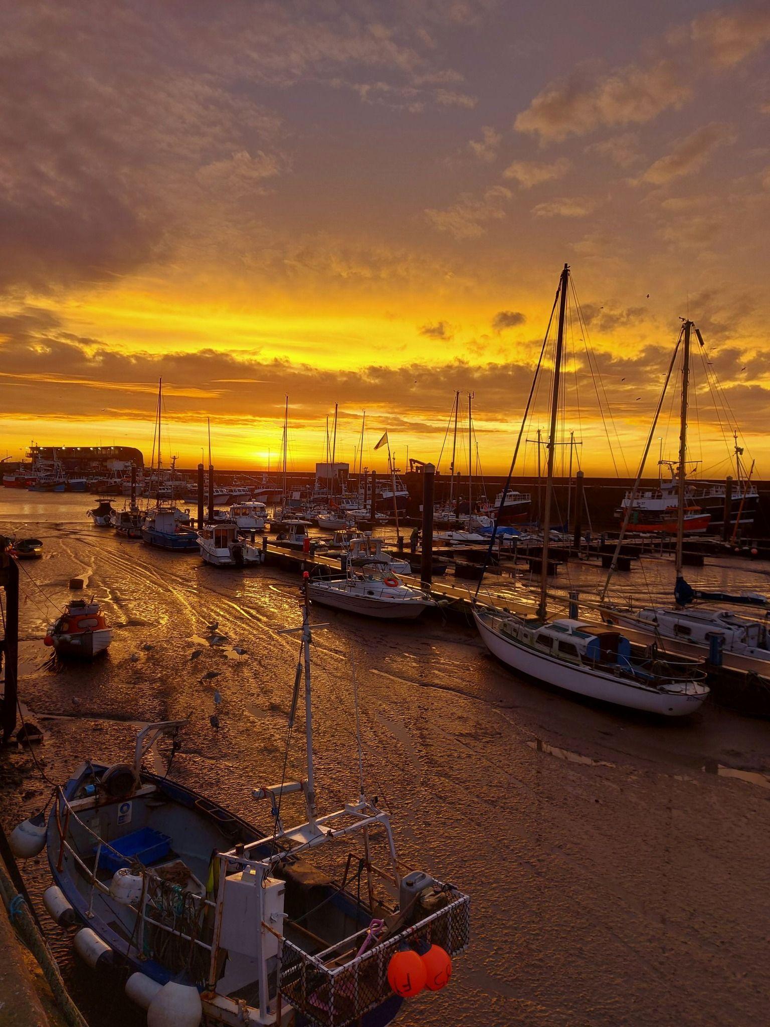 A photograph of lots of boats (20+) in the harbor, with a bright sky - a bright mix of oranges and yellows. The sailboats are nearly silhouetted but not quite - some detail can be seen - buoys, cargo. 