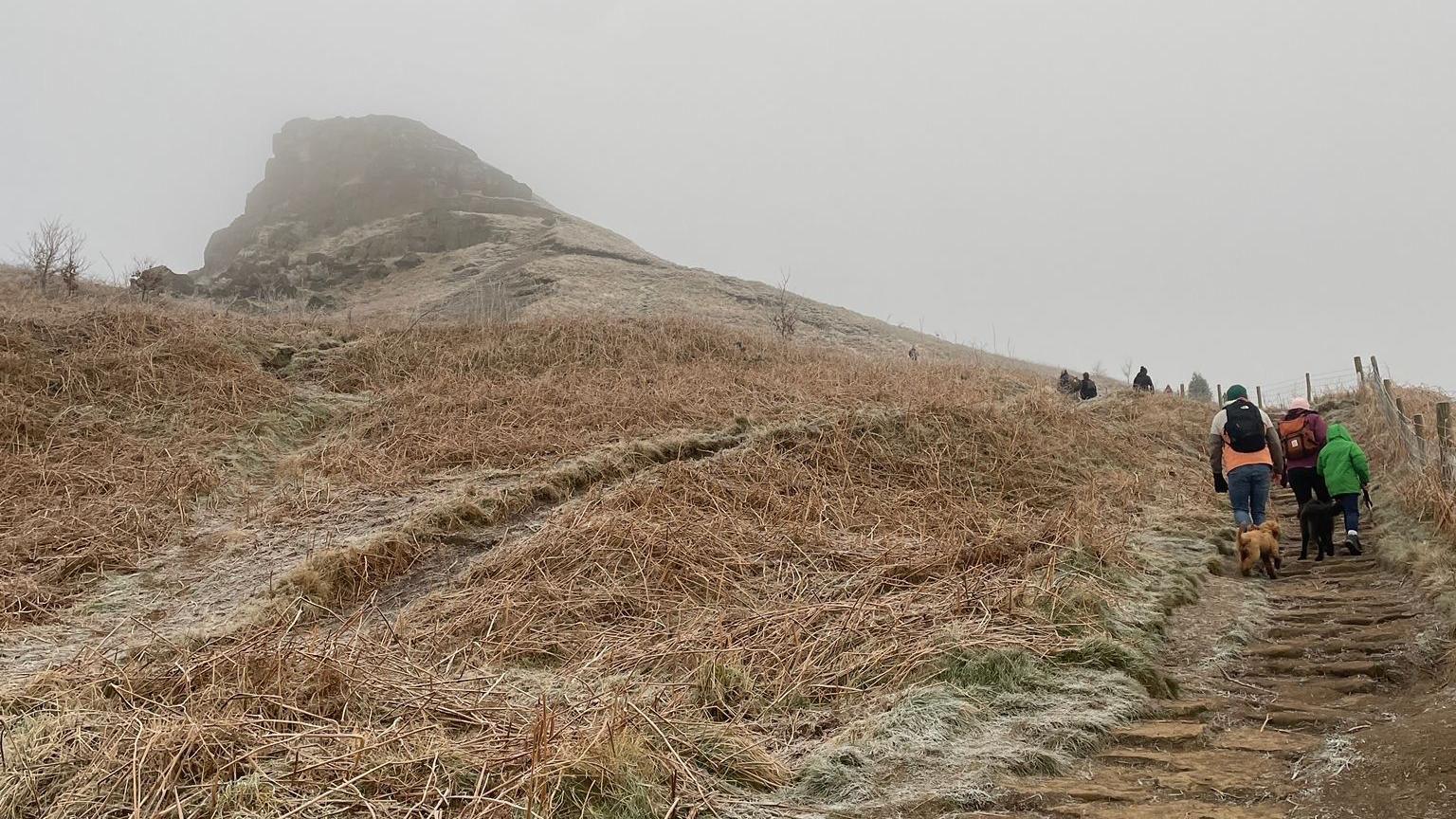 Roseberry Topping which is a large hill with frost-covered grass leading up to its peak. People can be seen walking up the stairs towards it.