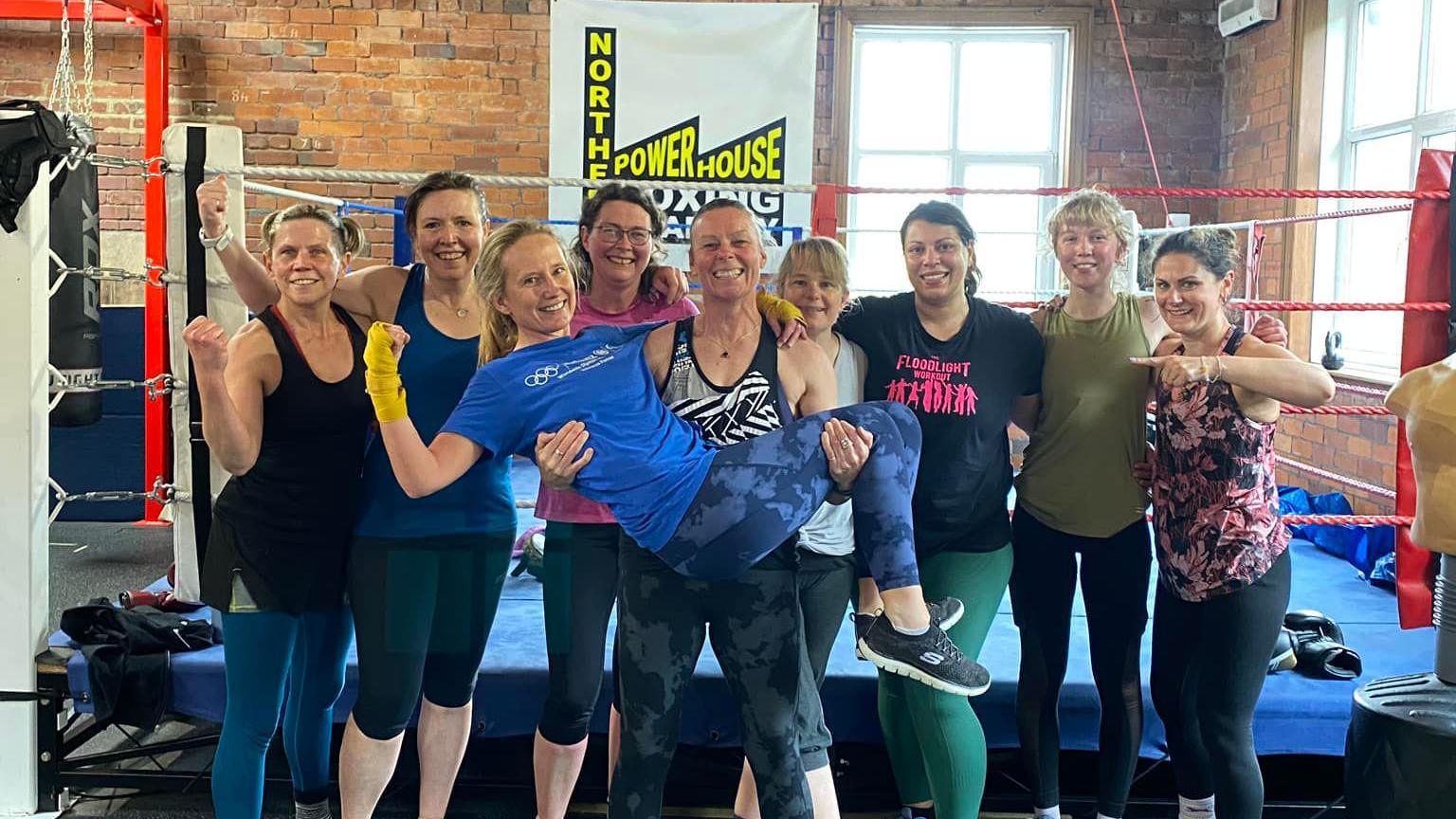 Nine women in gym gear stand in front of a boxing ring.