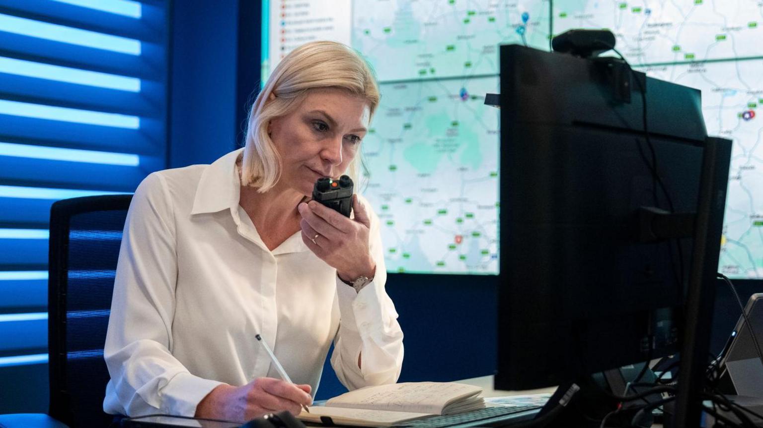 Georgina Bradley sat at a desk wearing a white shirt, holding a pen in her right hand with a book open in front of her and a walkie talkie in her left hand. There is a large map on a screen behind her.