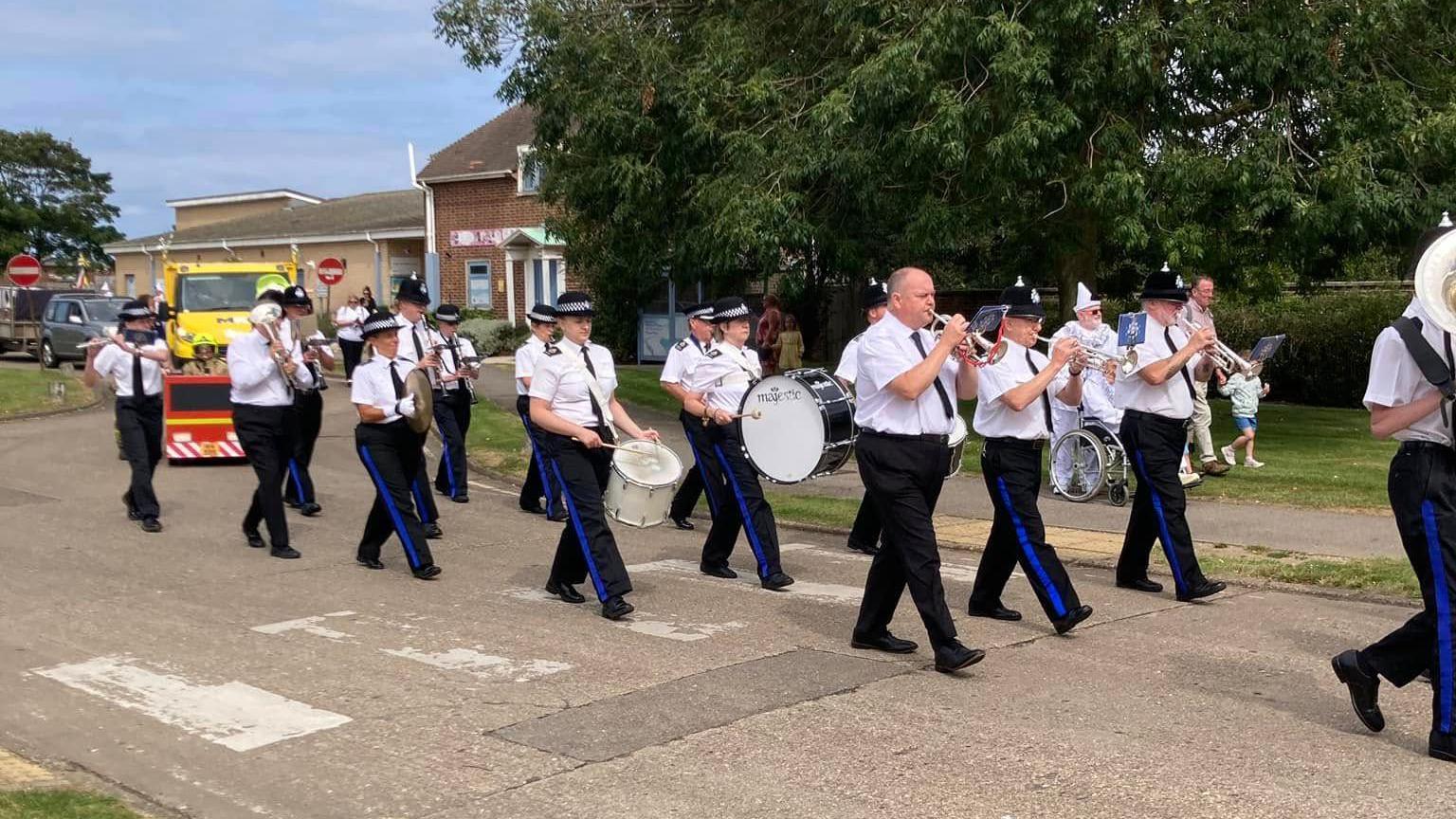 A marching band dressed in white shirts and dark trousers with blue stripes parades down a street. They are playing instruments. There is a yellow vehicle behind them. The sky is blue and cloudy.