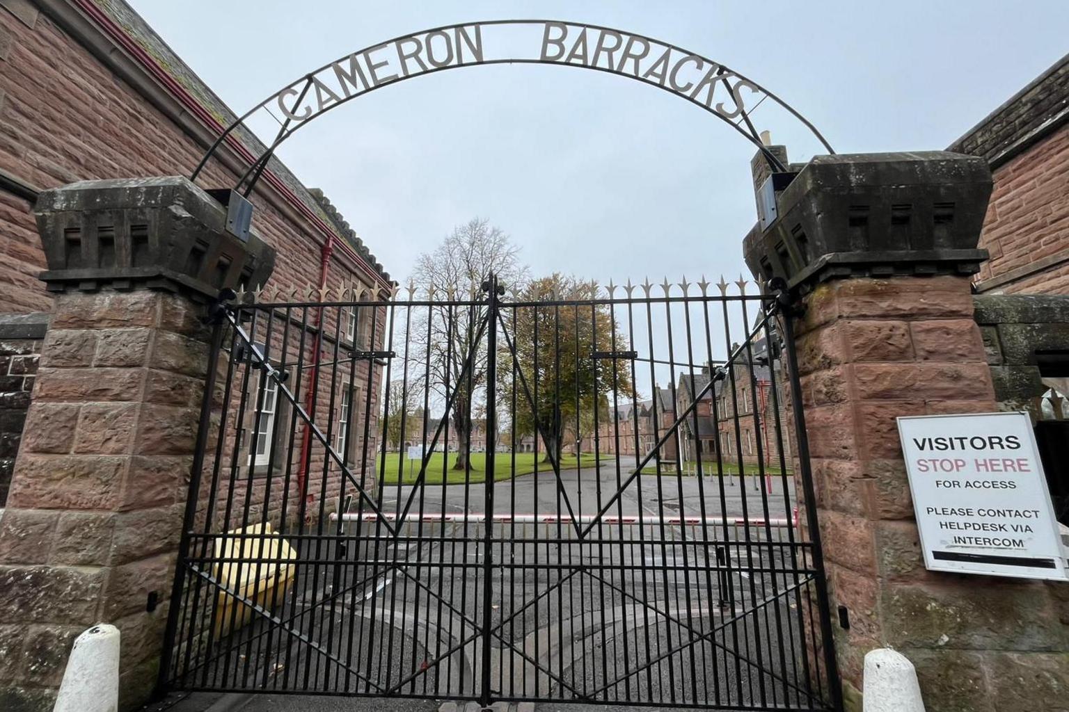 The barracks iron gates are shut. Above the gates is an arched metal sign which reads: "Cameron Barracks". The barrack buildings are built of red bricks.