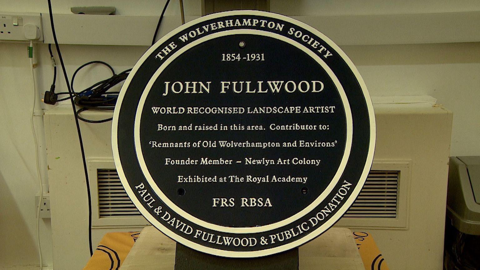 A round black plaque with white outlines, dedicated to artist John Fullwood