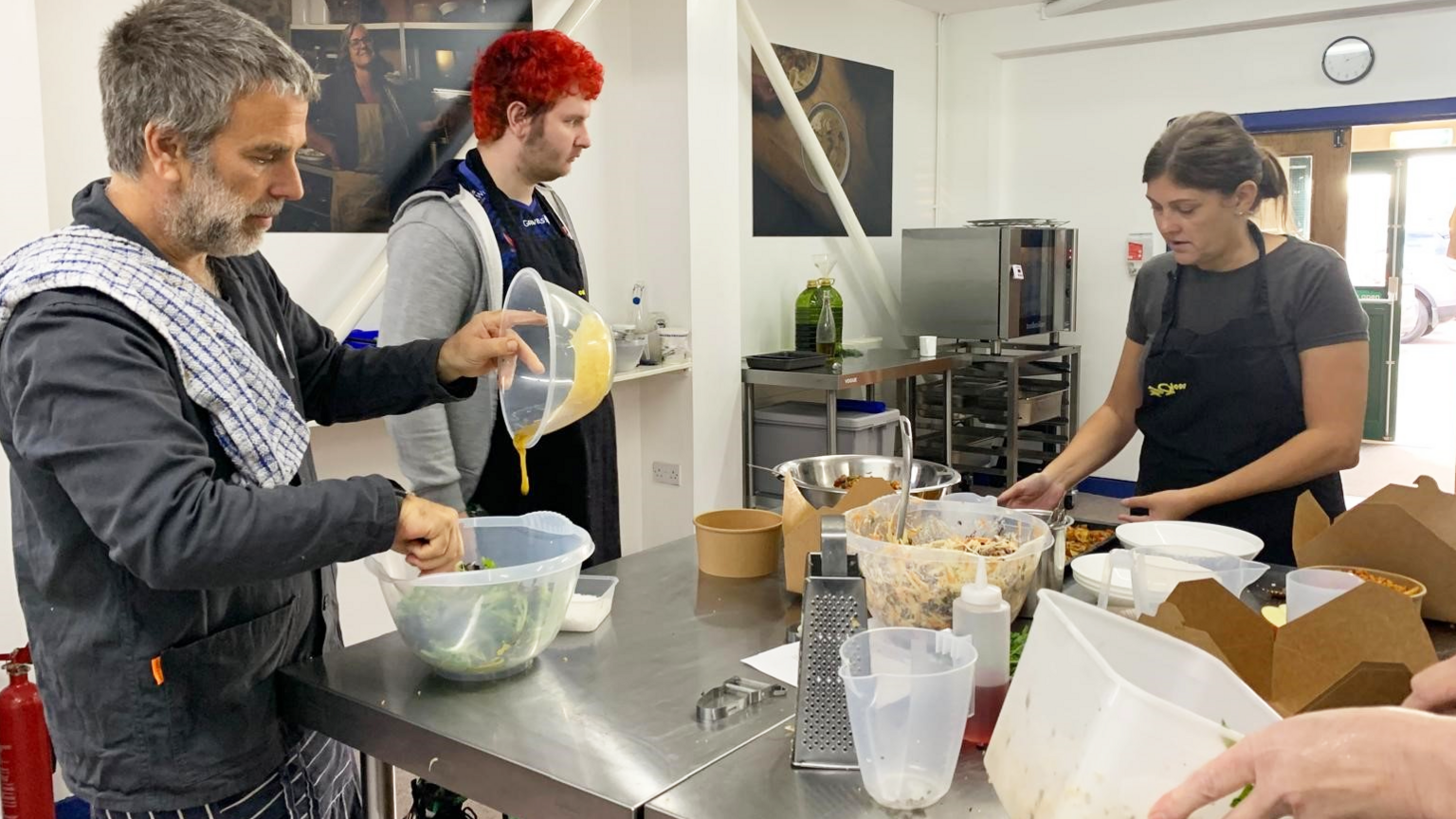 Lisa Davies and chef Tim Garrett in the Cook24 kitchen with others, preparing a meal on a stainless steel table top covered with food and cooking implements 