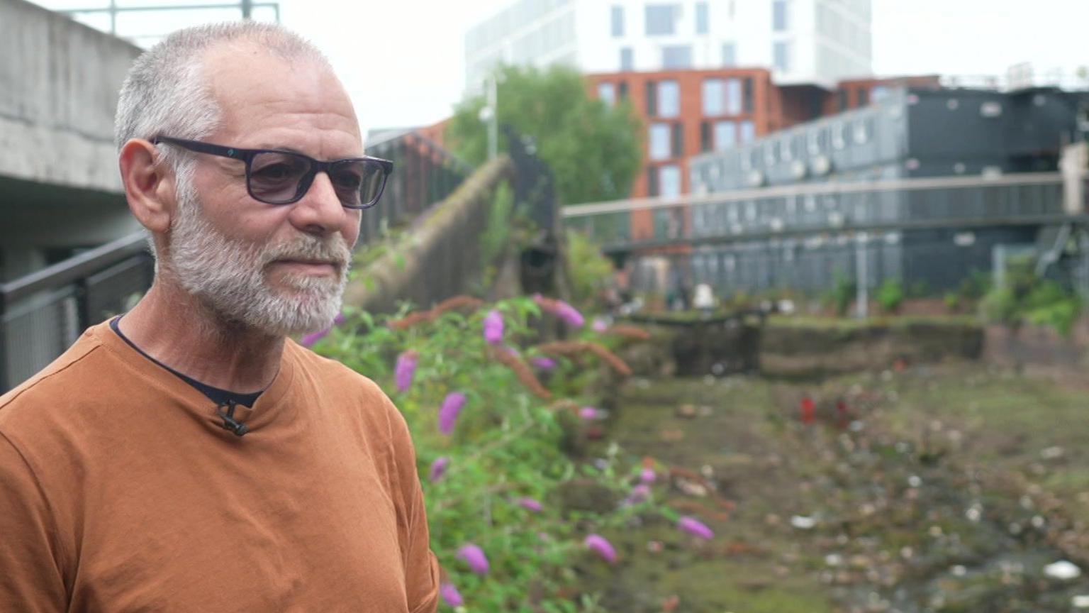 Marcus Brown, with grey hair and black sunglasses wearing an orange t-shirt, stands in front of a dry canal bed.
