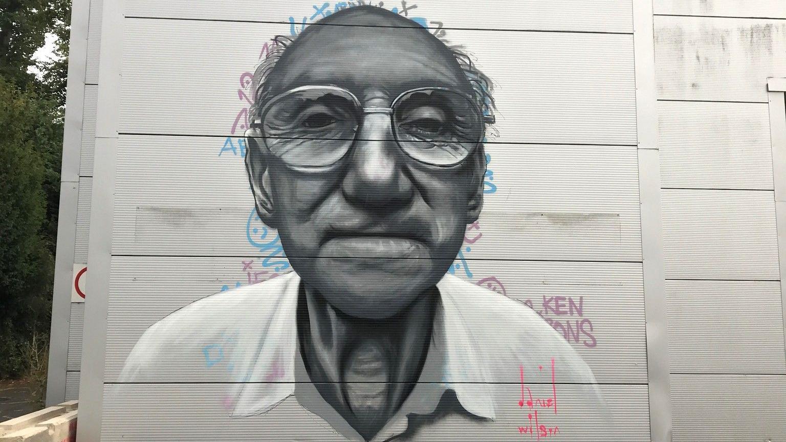 A mural of an elderly man, who has on glasses, looking straight on, painted on a building, with words behind the image and a signature at the bottom. There are trees to the left. 