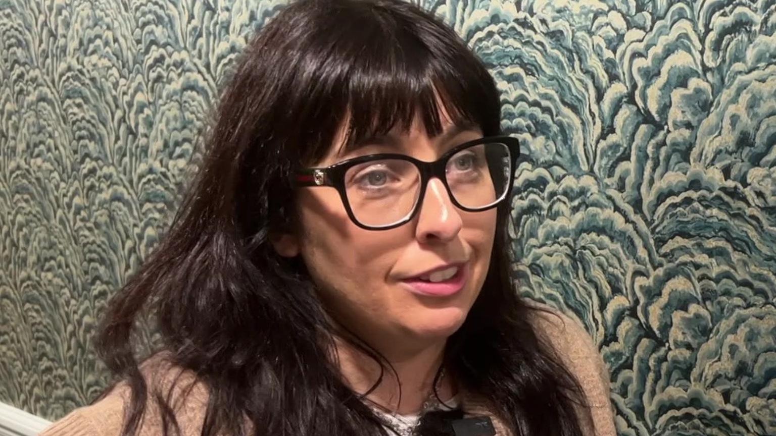Amy with long brown hair and glasses on in front of a patterned wall.