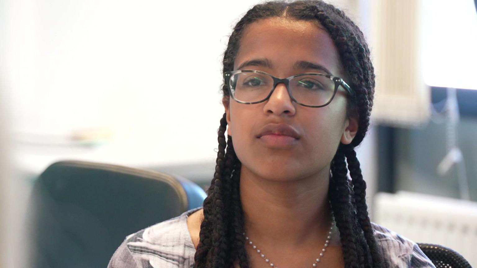 Alyssia, 17, is wearing glasses and has long braids. She is sitting at a table taking part in a discussion