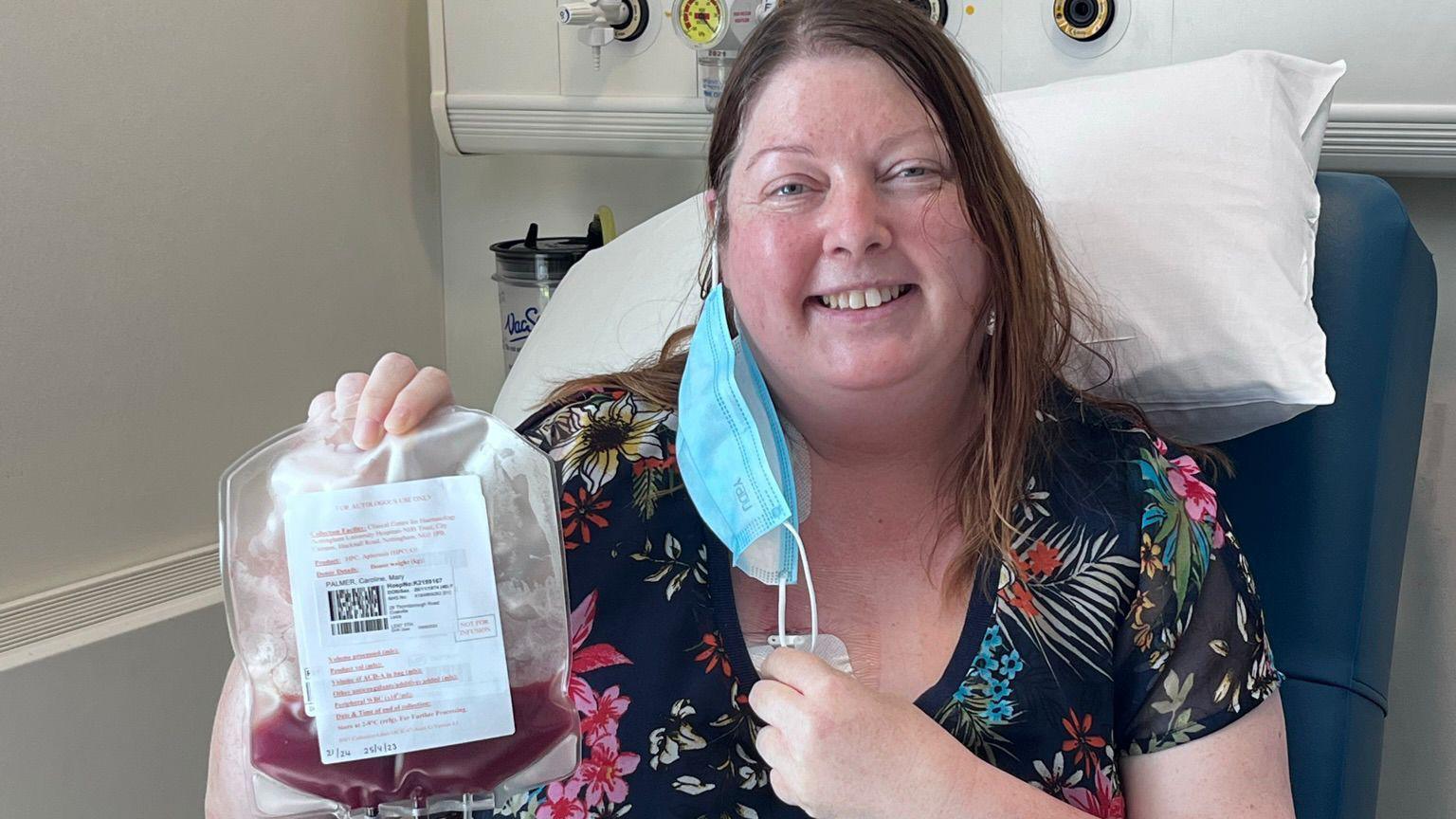 Caroline wearing a flowery top sat on a hospital chair. She has a blue face mask hanging from one ear and is holding up a bag of blood and smiling.
