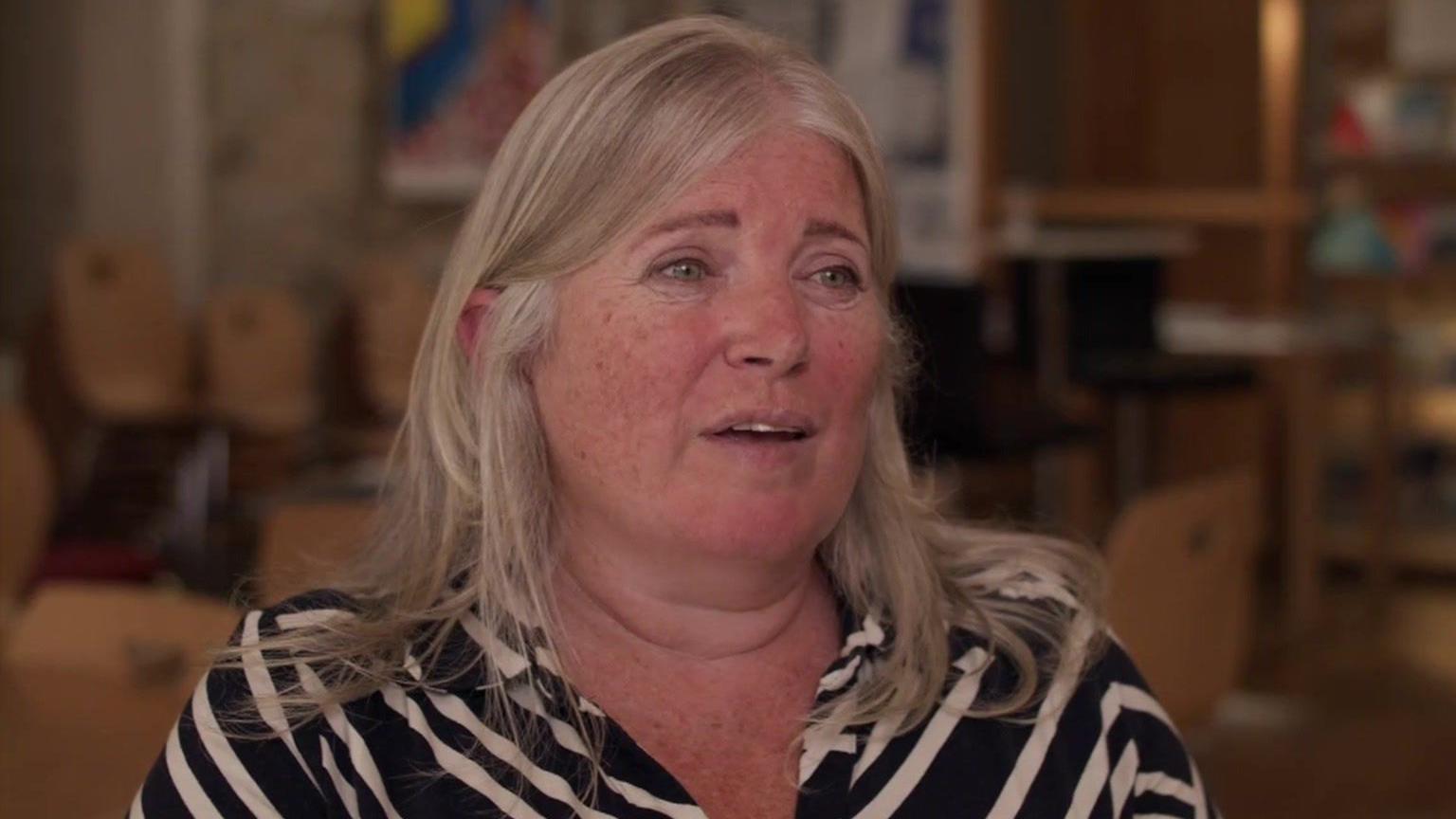 Professor Mackay has shoulder-length blonde hair and is wearing a shirt with diagonal black and cream stripes. The room behind her is blurred with a bookcase visible