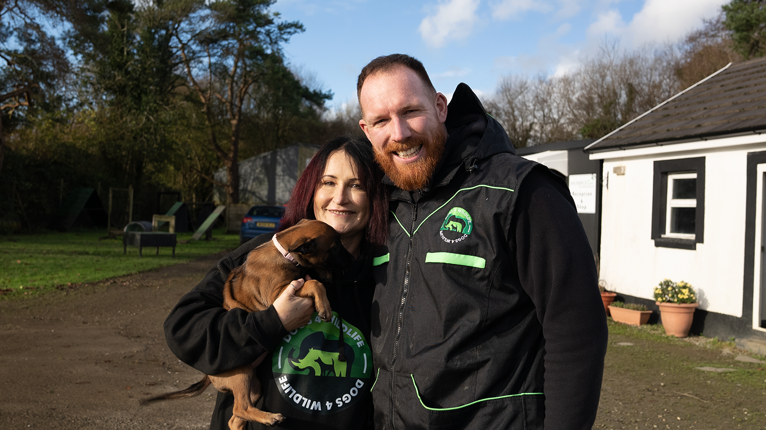 Jacqui and Drren wearing black jacket with their green logo which says Dogs4Wildlife. They are holding one of their Malinois puppies. To the right of them there is a white and black building and to the left of them there are trees and dog training equipment. 