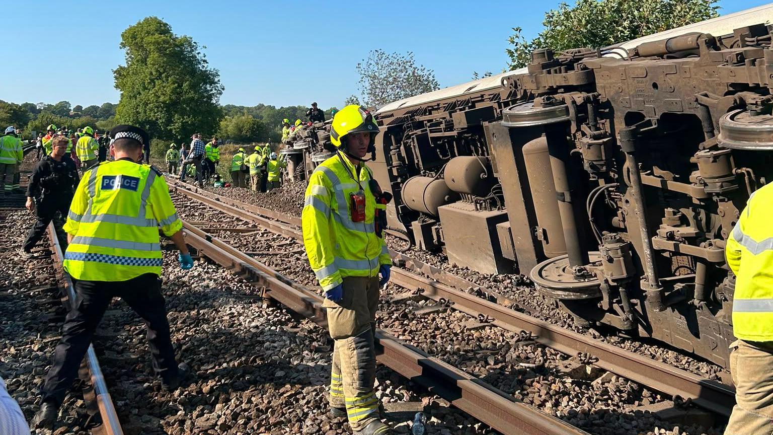 Emergency service workers in high vis jackets stand on the railway next to the train, which is overturned on its side