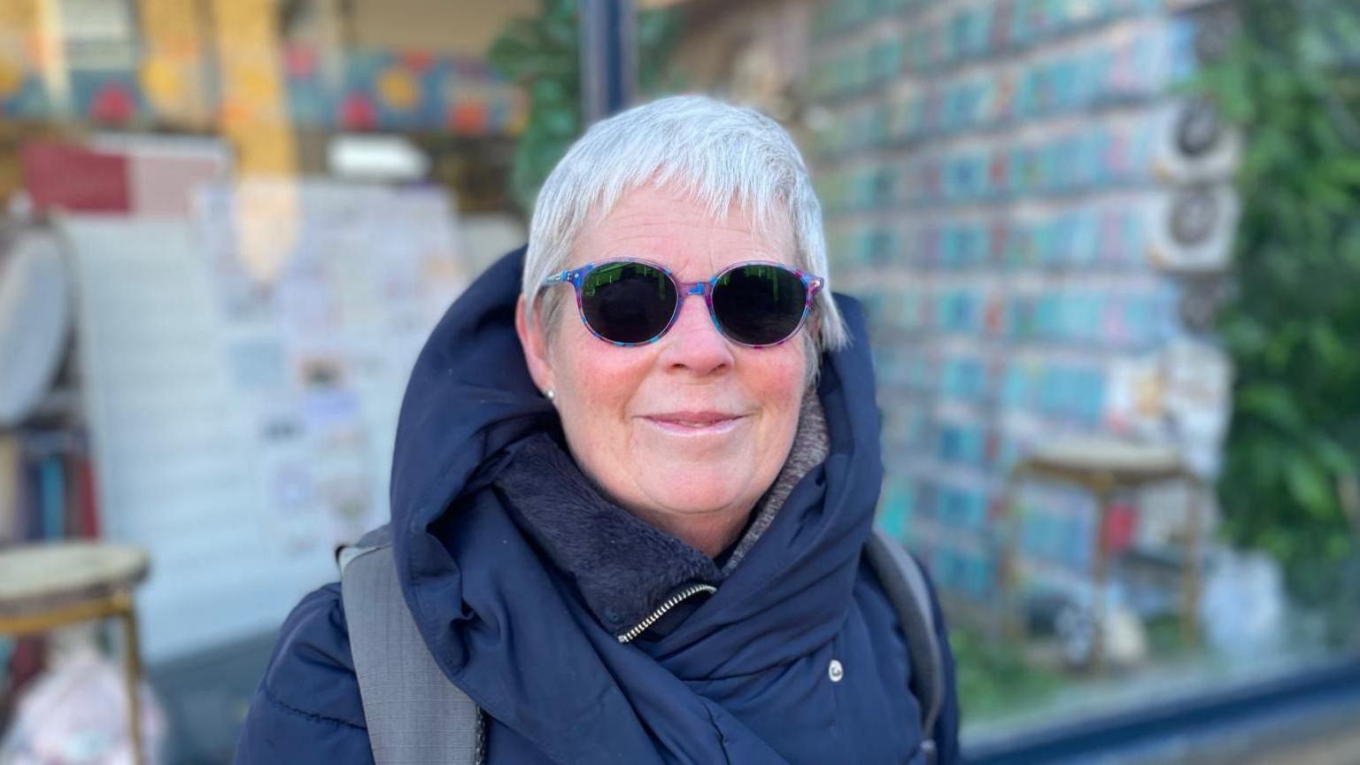 Lady with a blue coat on, and tinted glasses is standing infront of a shop in Elland.