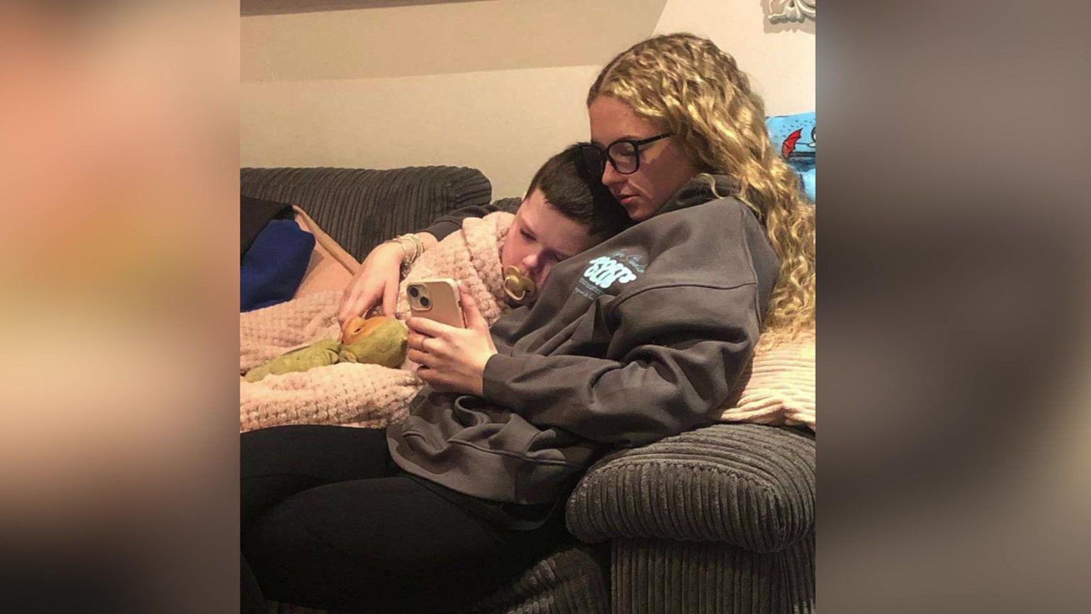 A teenager lies on a sofa with her younger brother. They are both watching something on a mobile phone.