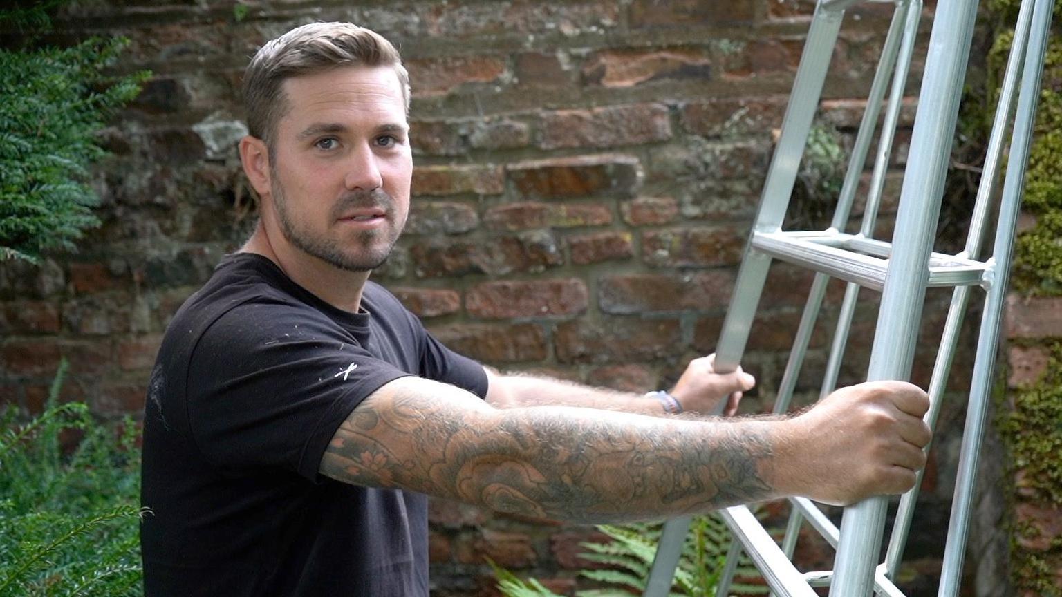 Andy stood at bottom of ladder he is holding while looking into camera 