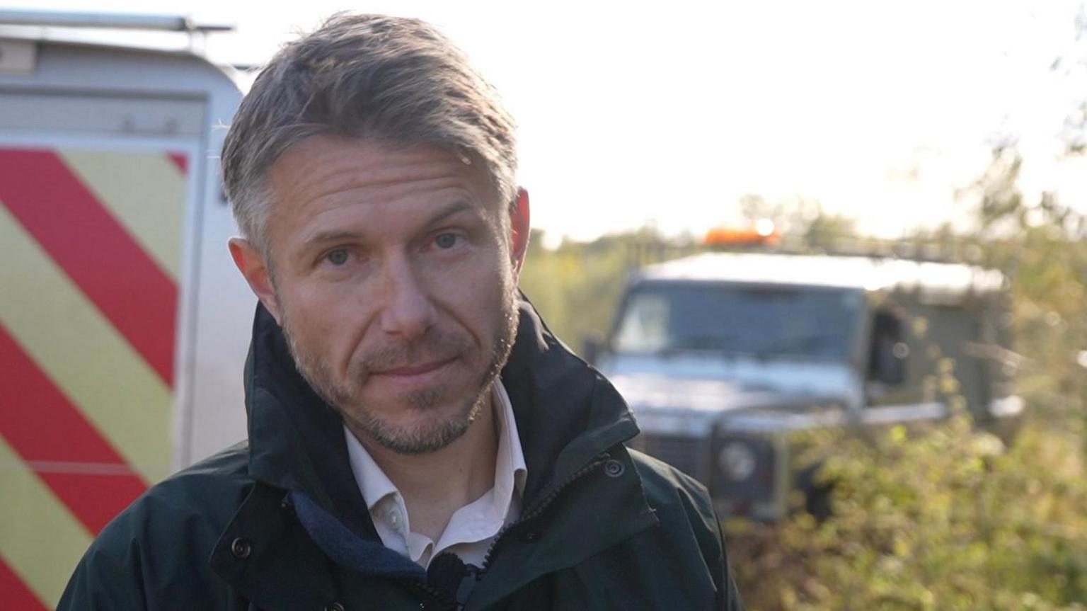 Aaron Dixey stands in front of two Environment Agency vehicles, which are out of focus. He has brown hair and a beard and is wearing a blue raincoat.