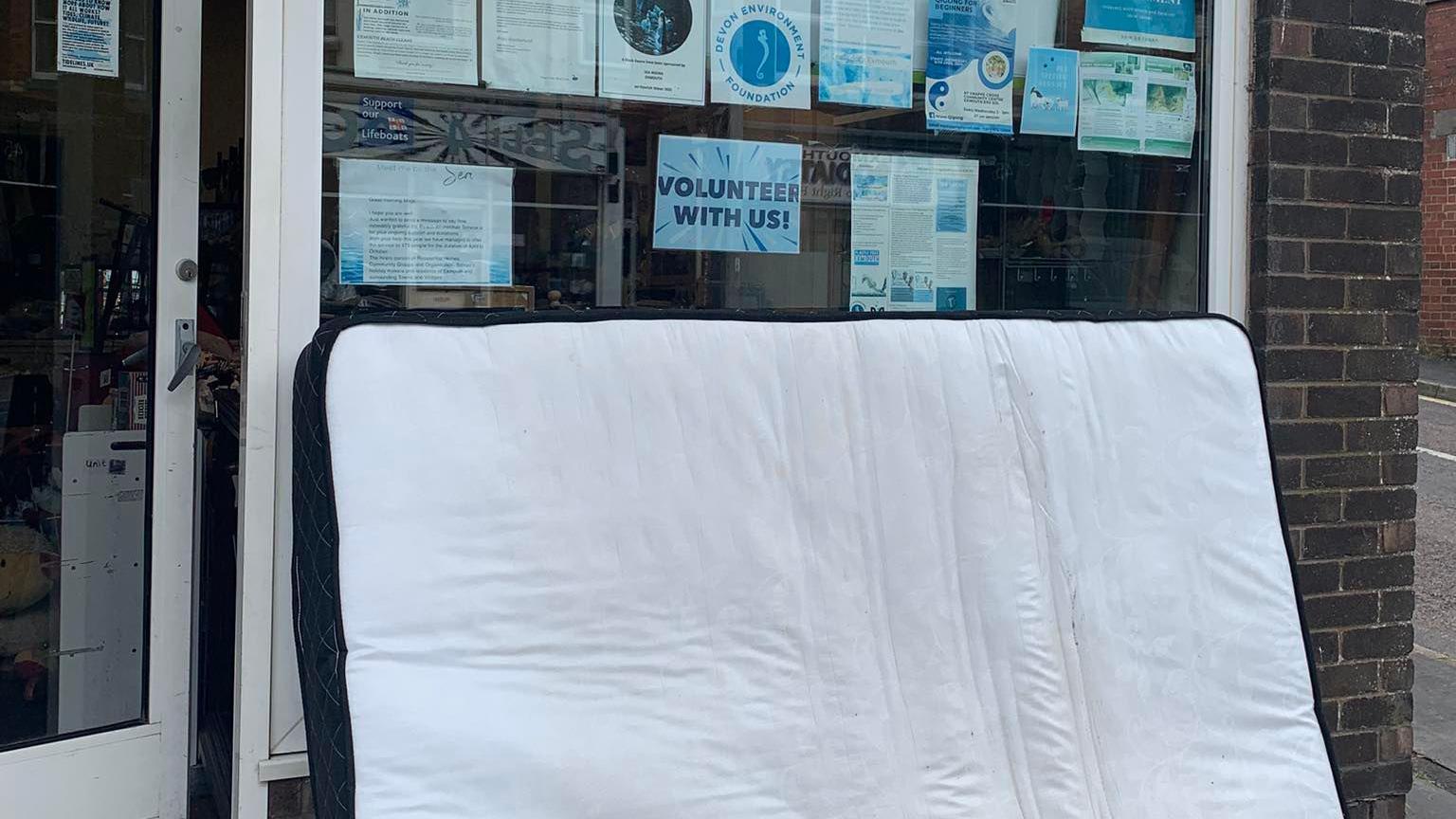 A large mattress fly-tipped outside of the charity shop. The mattress is white and there are signs/leaflets in the window. One of the signs says 'volunteer with us'. There is a door to the left.
