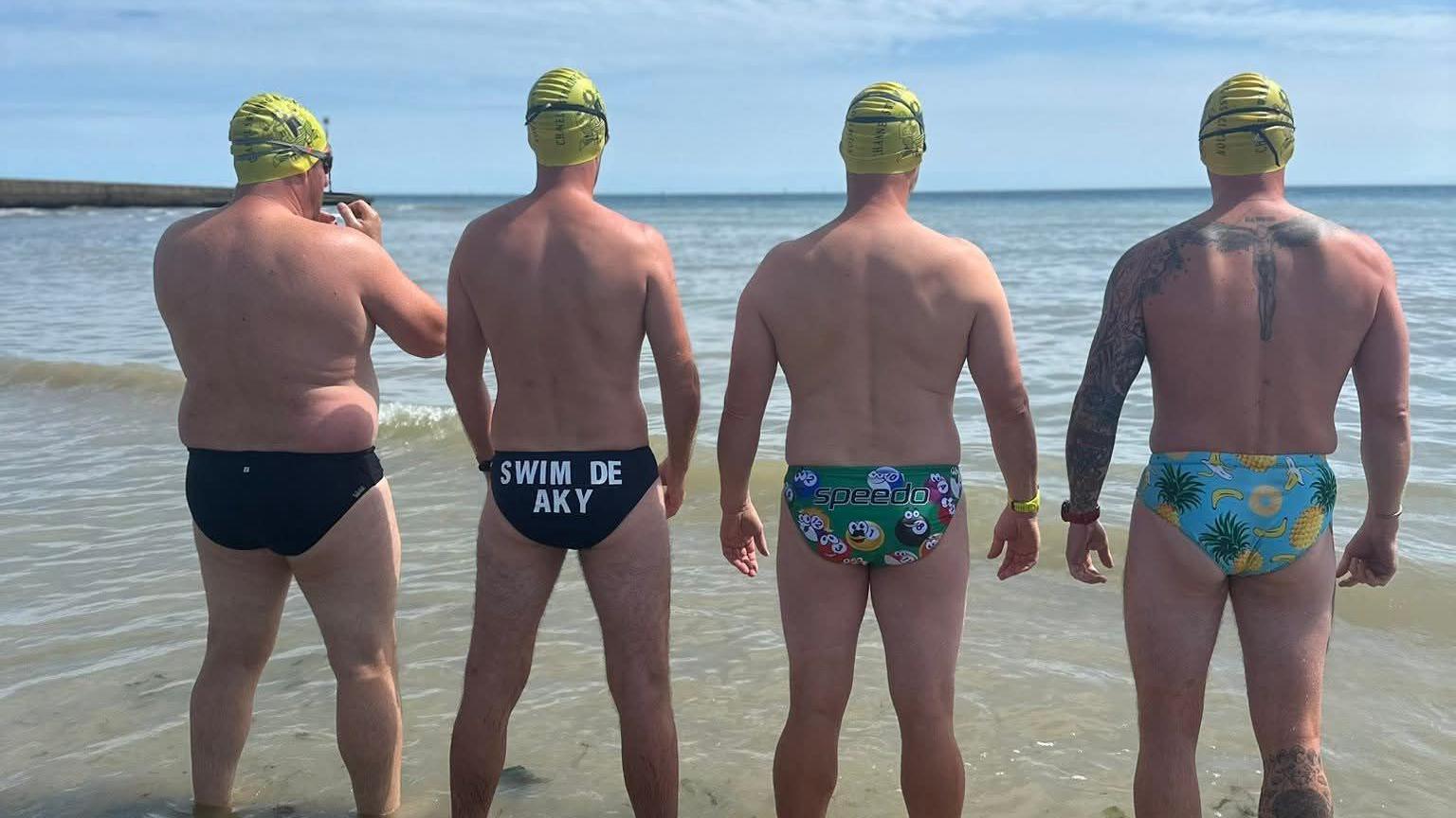 Four men standing in shallow water looking out to sea with their backs to the camera. All are wearing yellow swimming caps and speedo style swimming trunks.