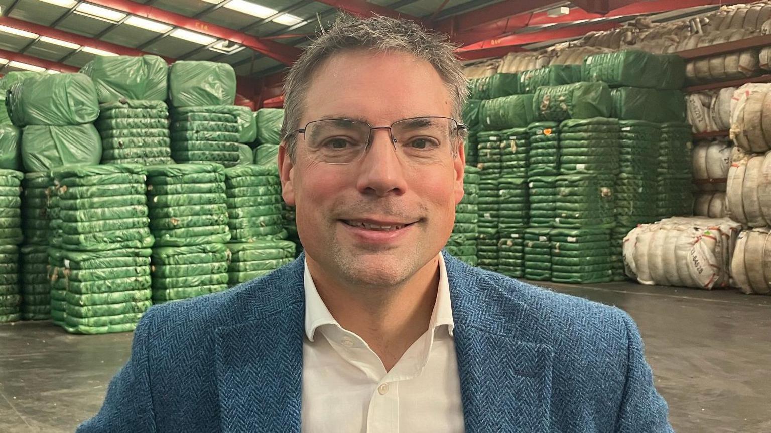 A man with short grey and brown hair and rectangular glasses stands in a warehouse. He is wearing a blue woollen jacket and white shirt.