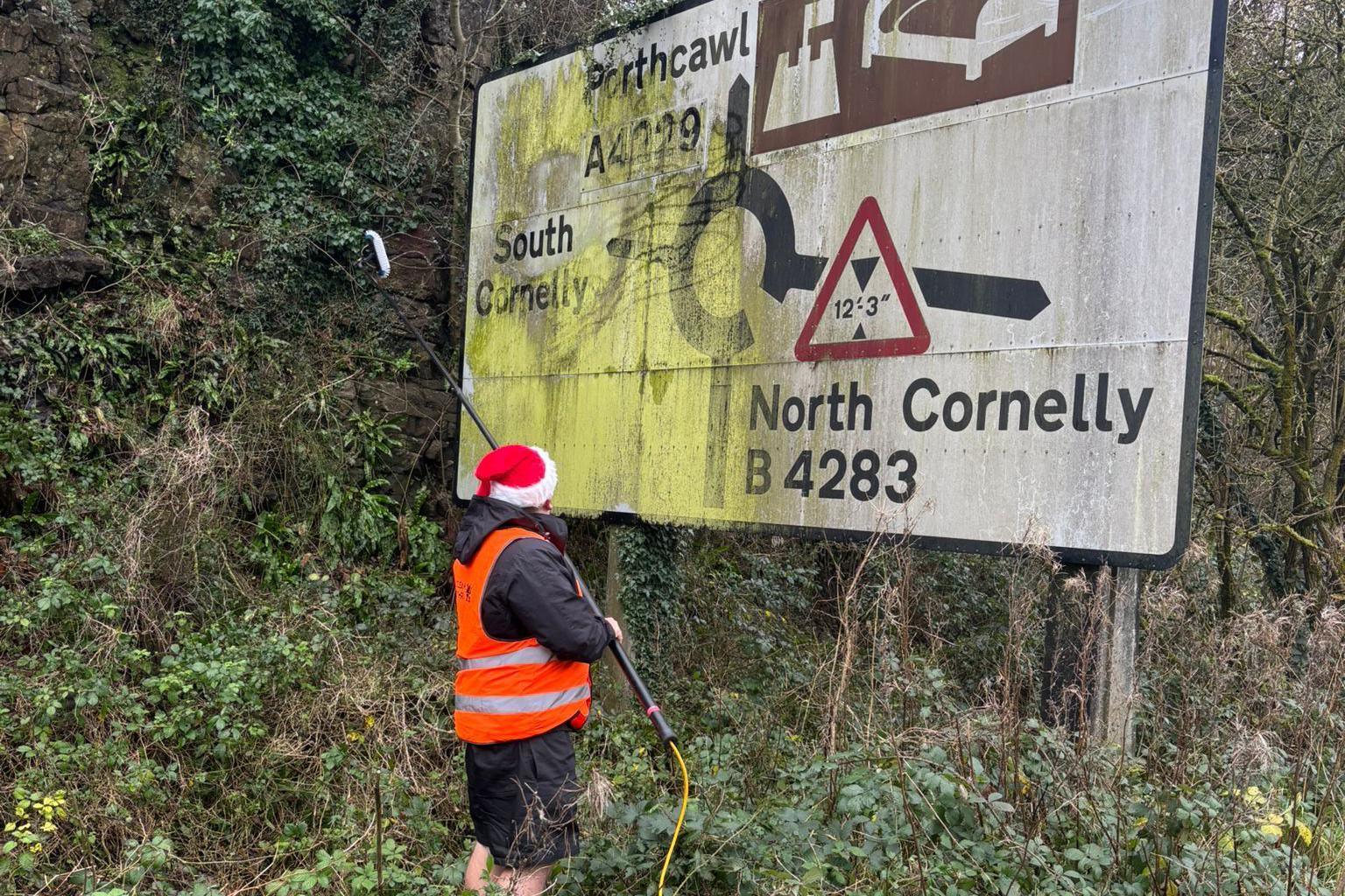 Man in front of a road sign cleaning it with window cleaning equipment. He is stood in bushes.