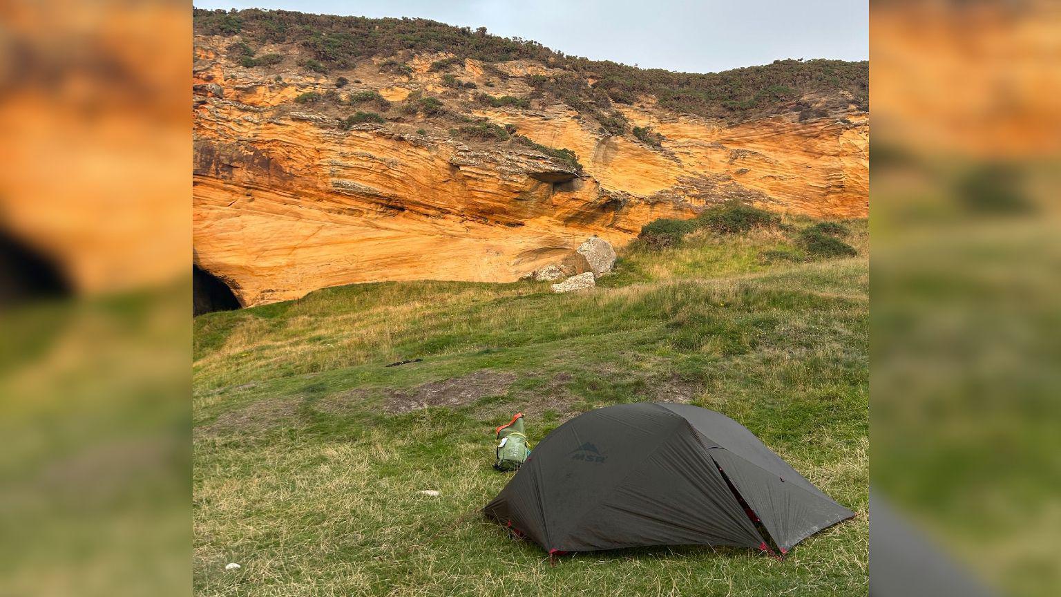 A grey one-man tent set up on a patch of grass beside a large rock cliff. It is sunset and the cliff is lit up orange