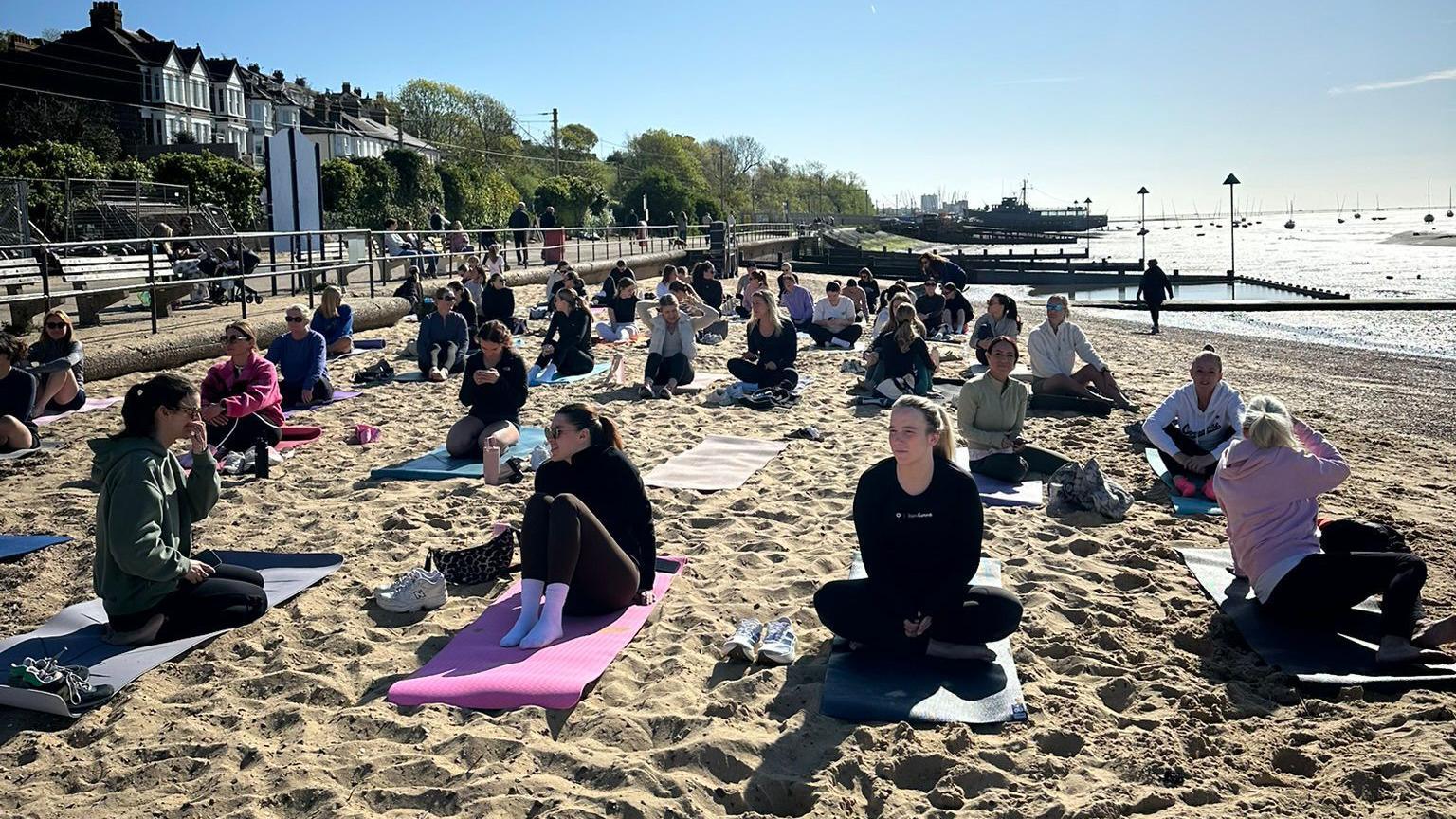 A dozens of people sitting on their yoga mats on a beach before or after a session, the sun is out