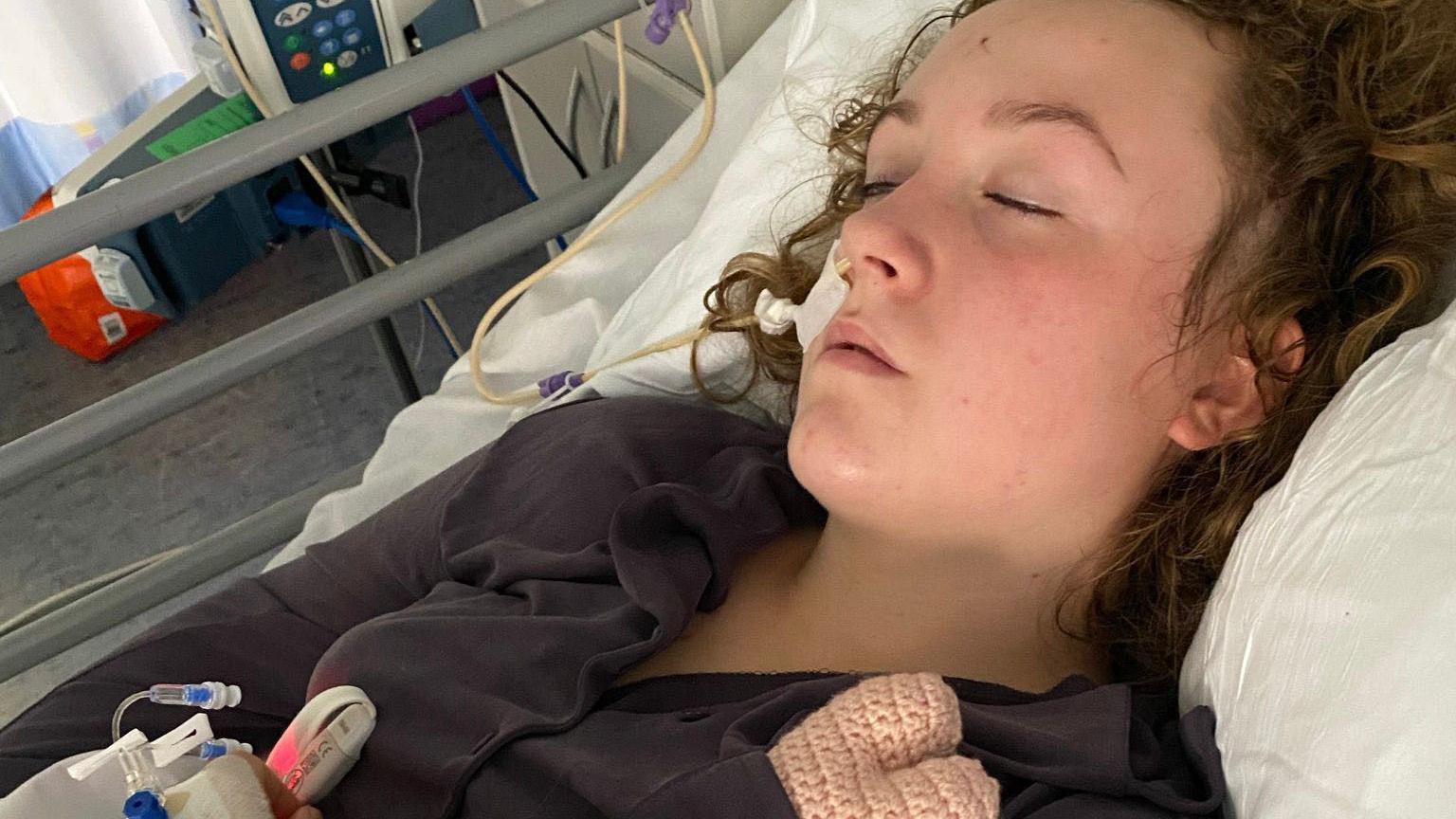 A young girl lies on a hospital bed with her eyes closed. Wires are attached to the side of her head.