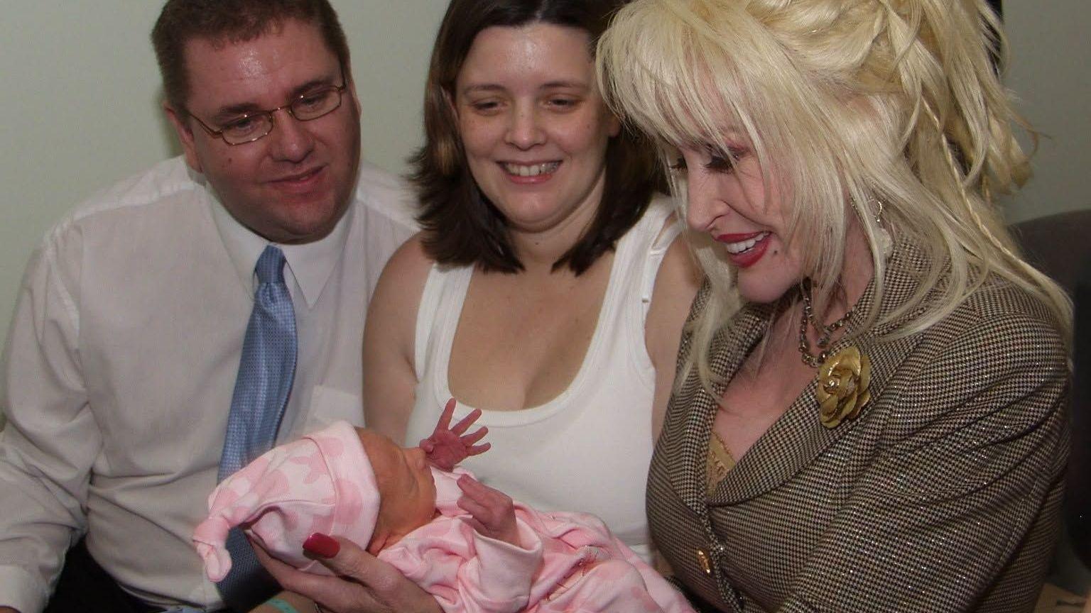 Three adults seated closely together indoors, focusing their attention on a newborn baby wrapped in a pink blanket.

In the foreground, Dolly is holding the baby carefully in both hands. She is wearing a light brown checked suit jacket with decorative gold buttons and a flower-shaped brooch on the lapel. The baby is dressed in pink clothing and a matching hat, with one small hand raised upward.

To the left of the image, Shane Jarman is seated wearing a white shirt, a light blue tie, and glasses. He appears to be looking down toward the baby. In the centre, Claire is seated wearing a white sleeveless top and is also looking toward the infant.