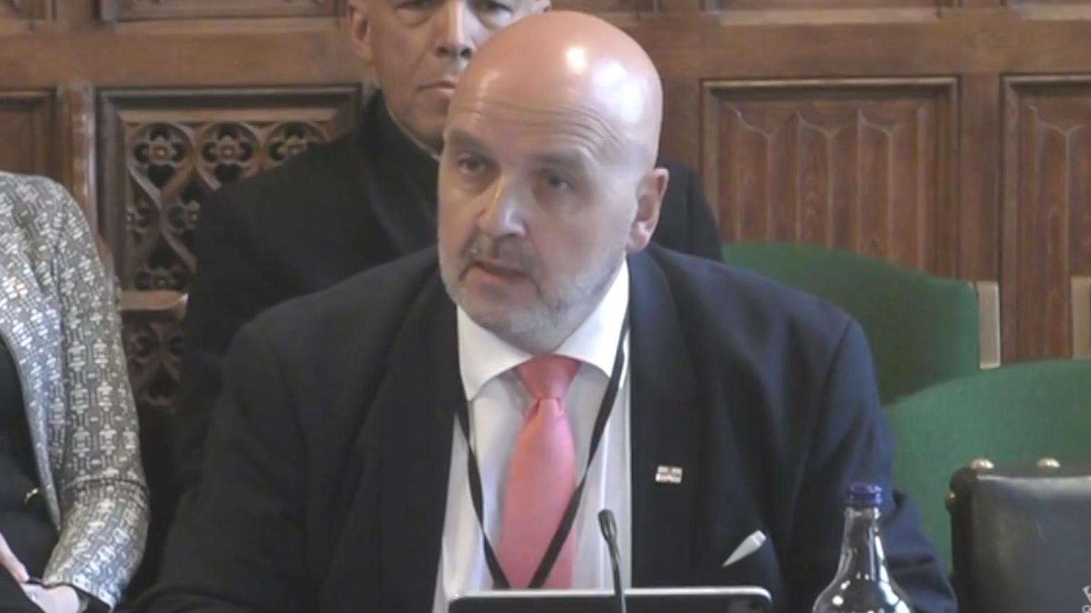 Deputy Jonathan le Tocq in the UK House of Parliament - He is a bald man wearing a blue suit, a white shirt and a pink tie. On his left lapel he is wearing a Guernsey flag pin badge.