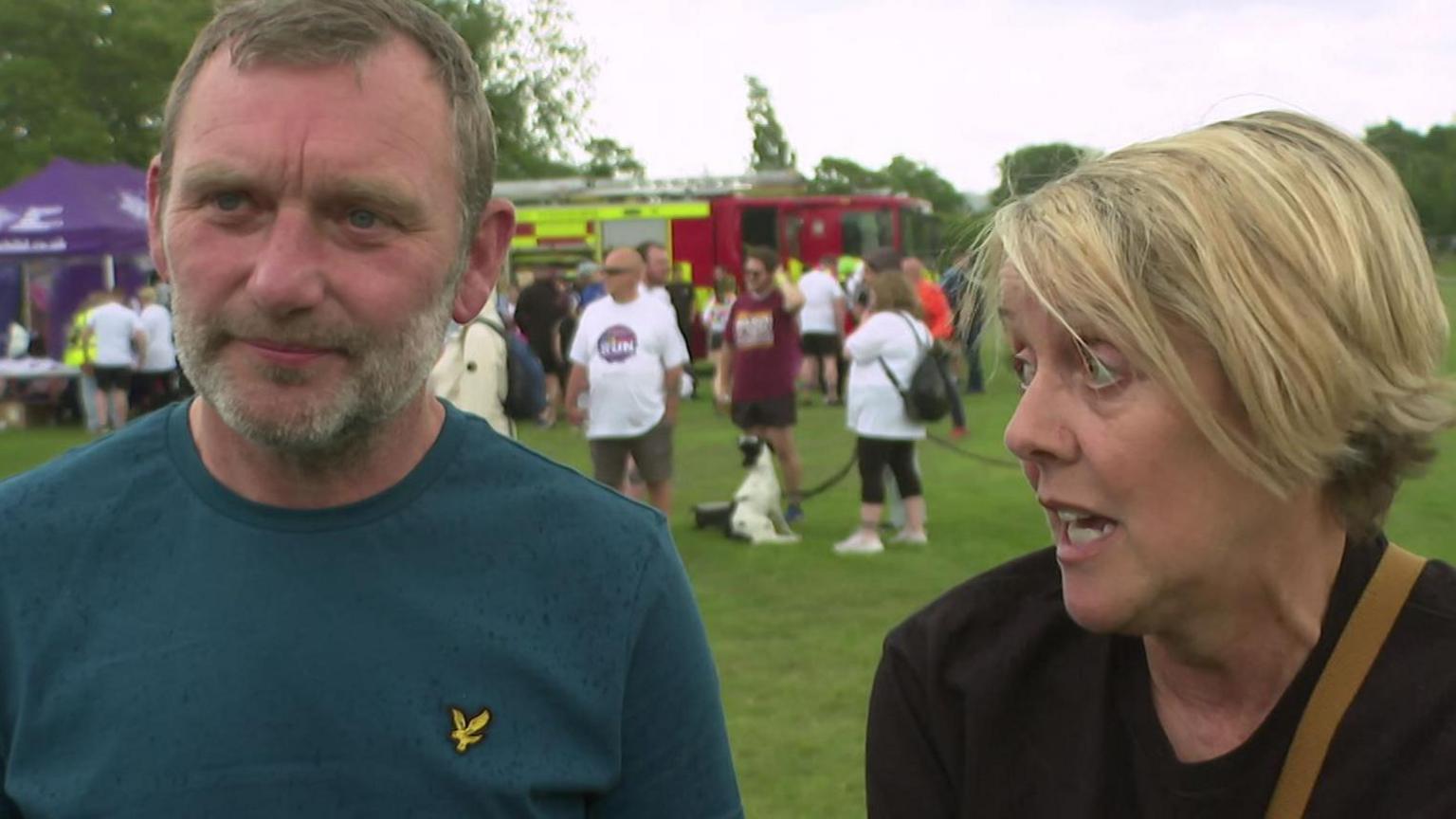 A red faced man with greying hair and a beard next to a blonde woman wearing a black top. In the background are fun runners and a fire engine.