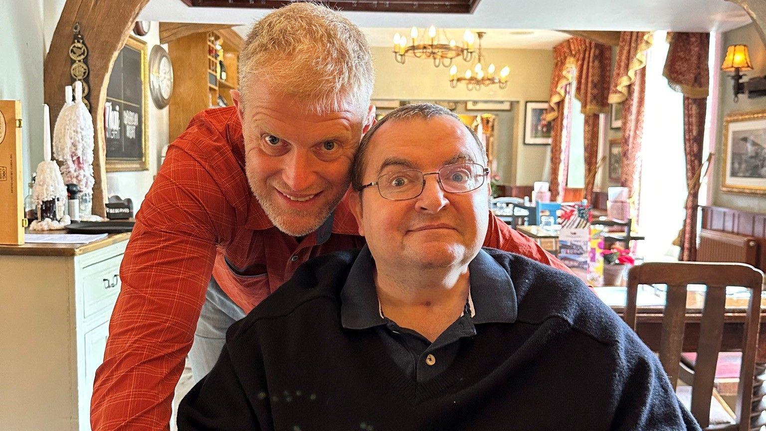 A man sitting down wearing glasses and another man bending over him smile at the camera with a restaurant interior behind them