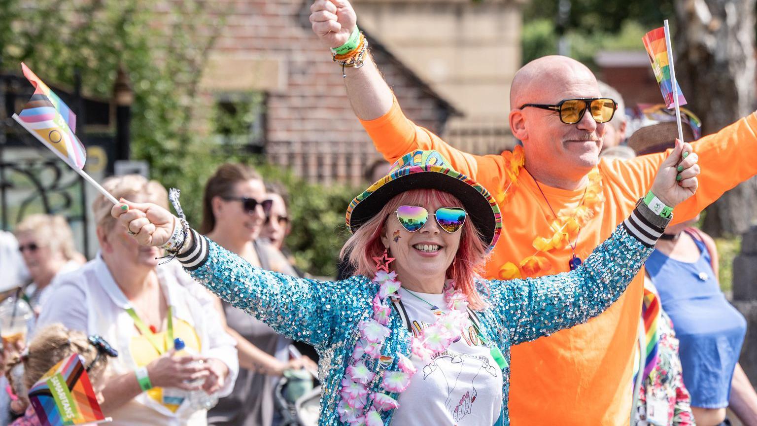 A woman in heart shaped glasses, rainbow coloured hat and a blue sequinned jacket holds rainbow flags aloft. Behind her is a man in an orange t-shirt and floral garden, also waving flags. They are walking in a parade and others in colourful clothing waving flags can be see behind.