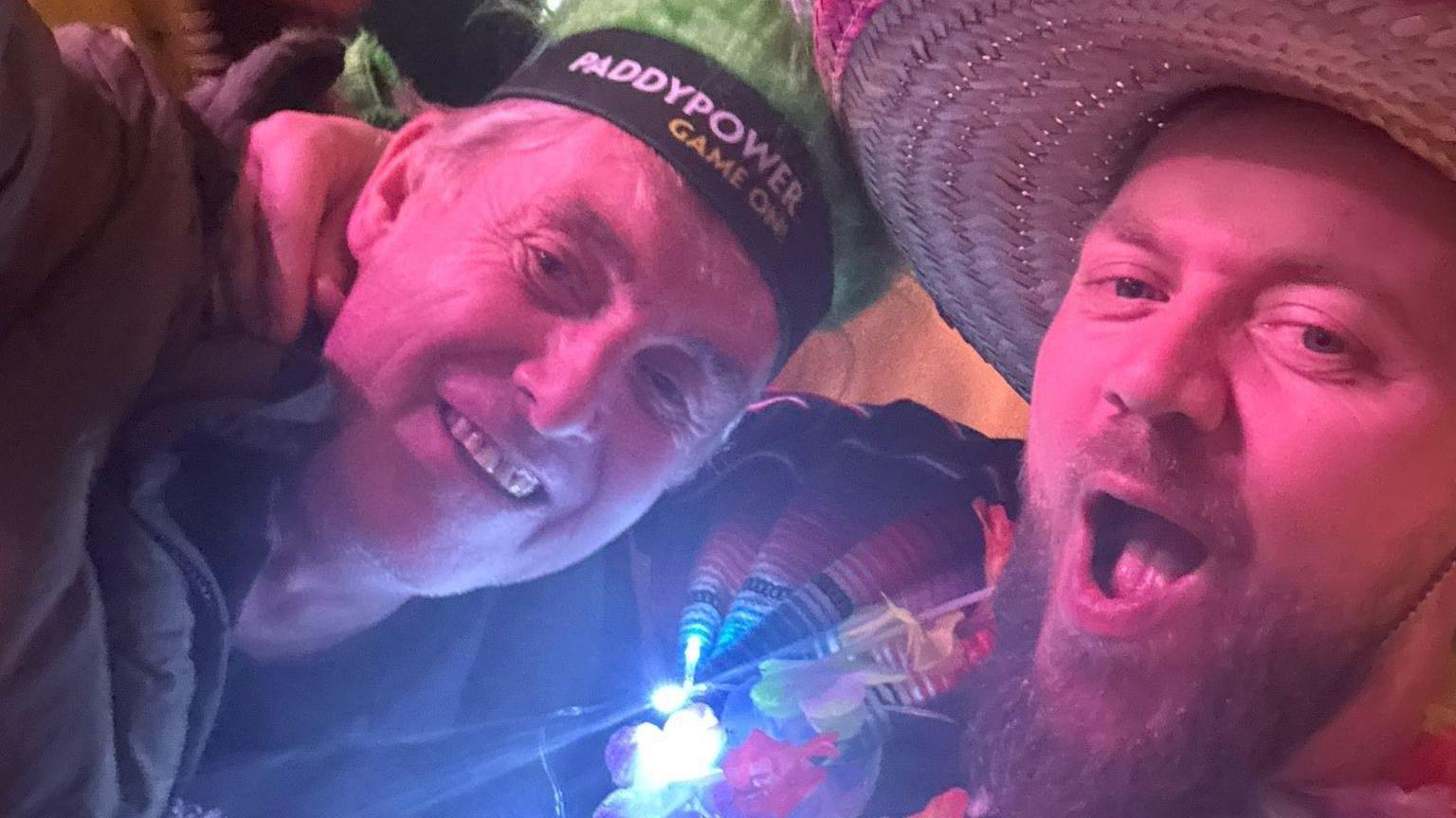 Craig Wells, who wears a sombrero, has his arm around his homeless guest John in a selfie. John is older and wears Paddy Power headgear. Both men are smiling.